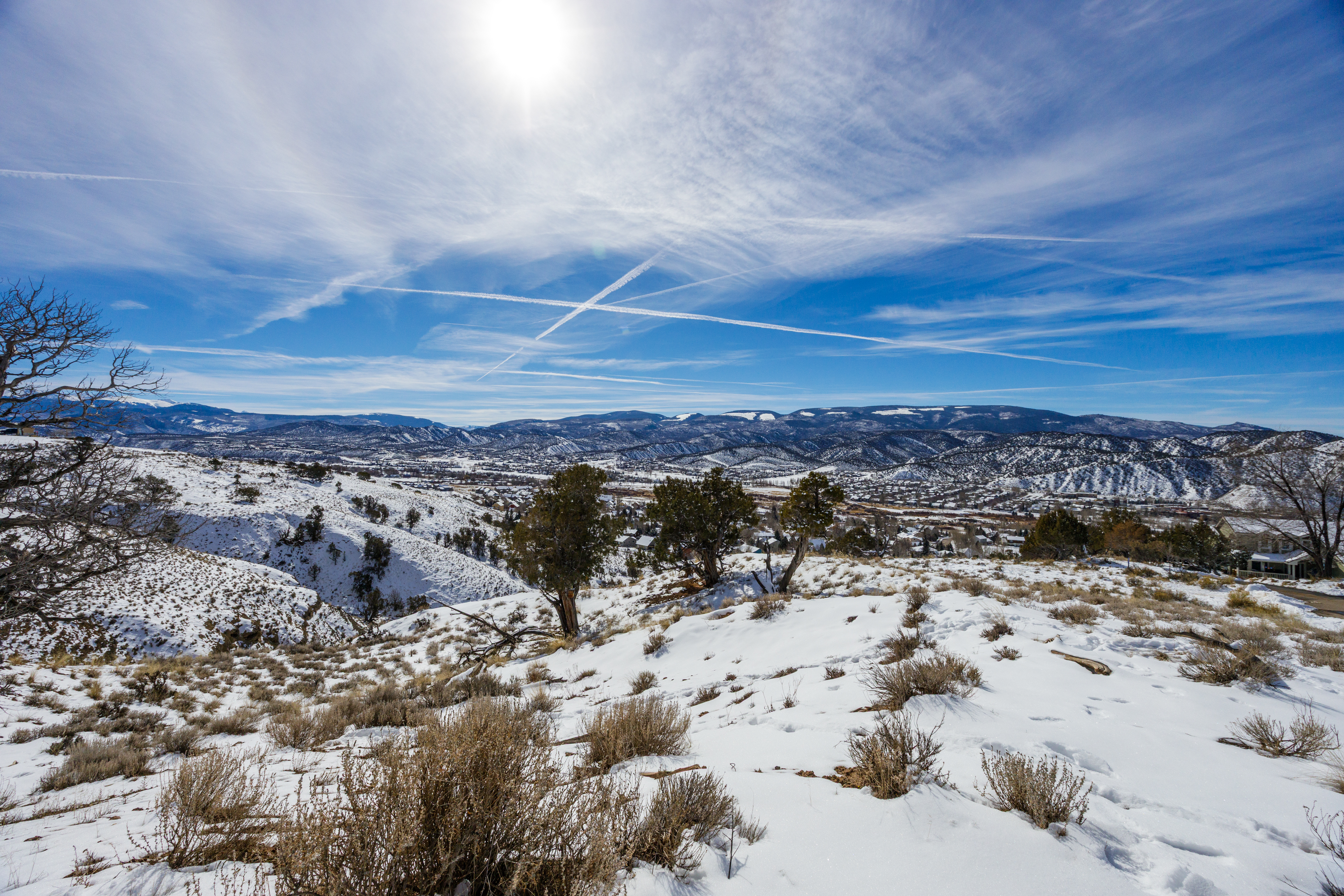 This aerial view showcases a snow-covered landscape with rolling hills and distant mountains. The sky is bright blue with wispy clouds and contrails, adding to the serene yet dynamic atmosphere. The landscape hints at a tranquil and secluded property location, appealing to potential buyers seeking privacy and natural beauty.