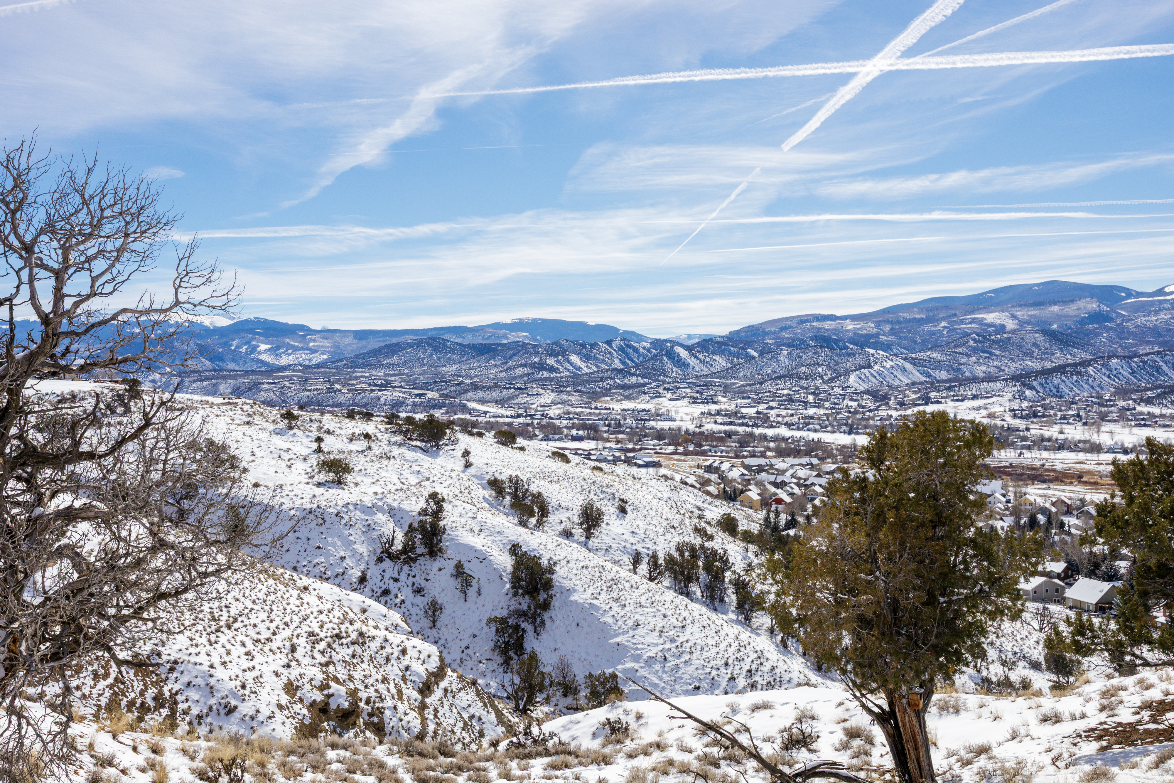 This image shows an aerial view of a snow-covered town nestled in a mountain valley. The landscape features rolling hills with scattered trees and houses, all dusted with snow. The sky is filled with contrails, adding a sense of depth and dimension to the vast landscape.