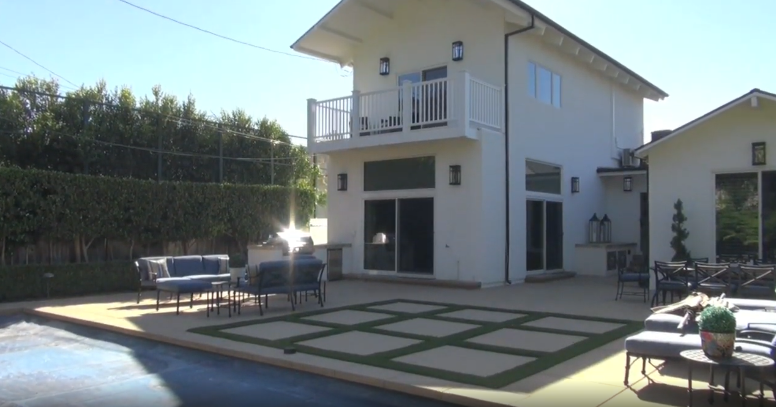 This bright, contemporary rear exterior showcases a multi-level layout with a second-story balcony and large sliding glass doors leading out to a paved patio area. The outdoor living space is enhanced by well-arranged patio furniture, a swimming pool with a cover, and a creative grid-patterned paver design set against artificial turf. It is a highly functional and modern backyard space ideal for entertaining and outdoor leisure.
