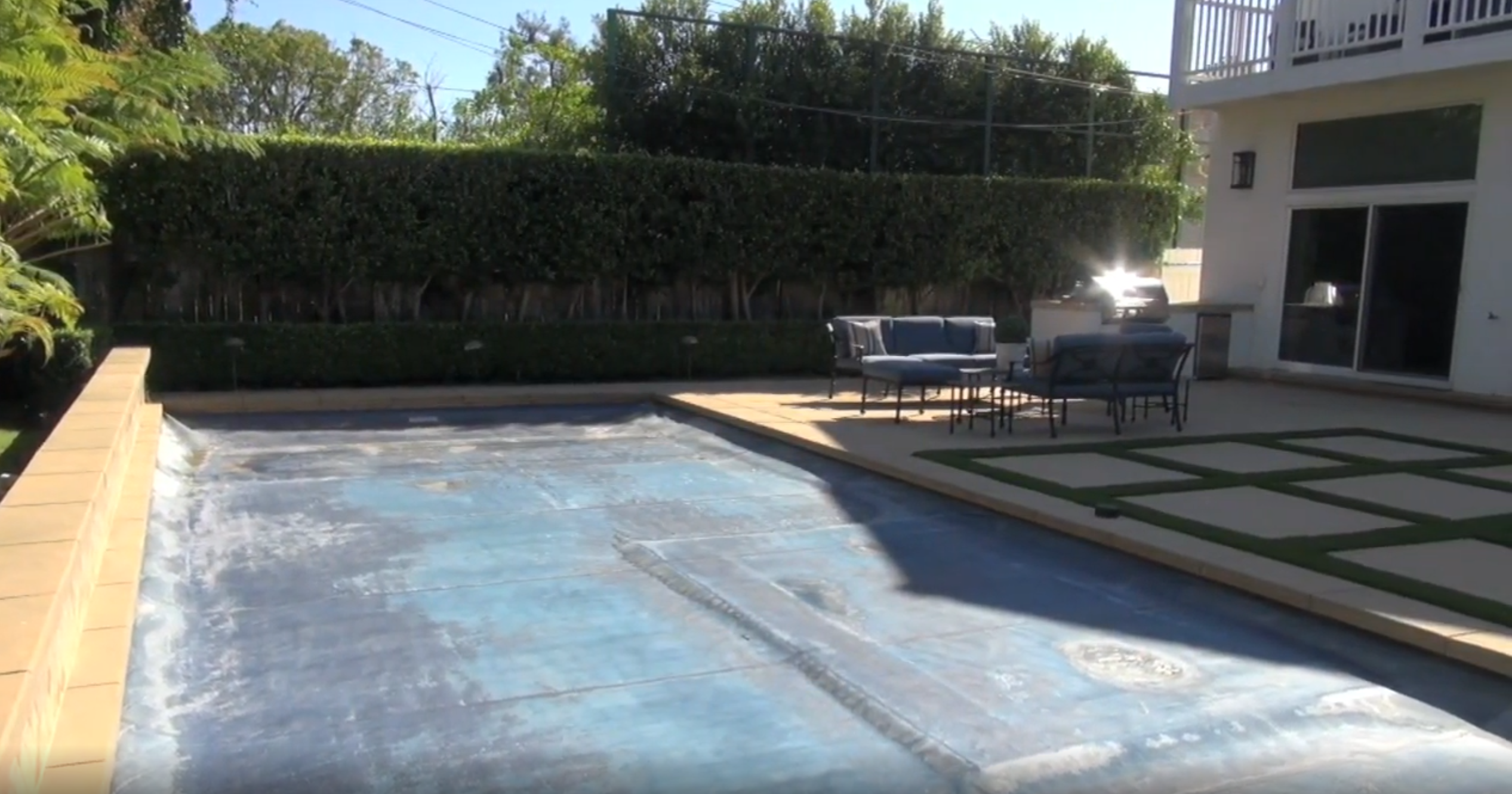 This image features a backyard oasis centered around a swimming pool equipped with a safety cover. Adjacent to the pool is a concrete patio area furnished with outdoor seating, complemented by a patch of modern square pavers set in artificial turf. The space is private and well-defined by tall, manicured hedges, making it an ideal setting for outdoor entertaining.