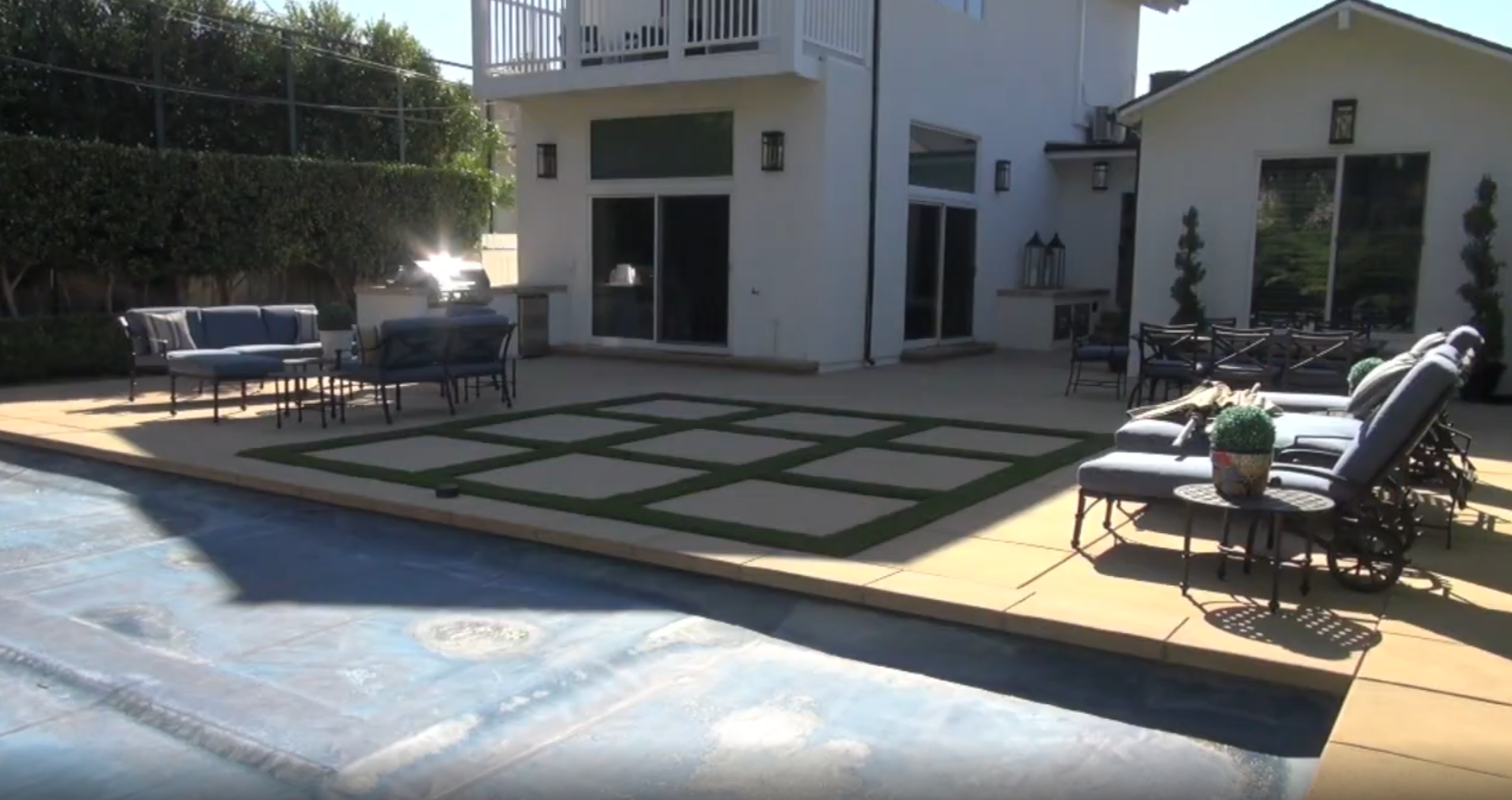 This spacious backyard patio area features a covered swimming pool, comfortable outdoor seating arrangements, and a unique grid-patterned paver design with integrated artificial turf. The space is adjacent to the home, creating an inviting transition between interior and exterior living areas. With its clean lines and functional layout, the area serves as an ideal setting for outdoor dining and leisure.