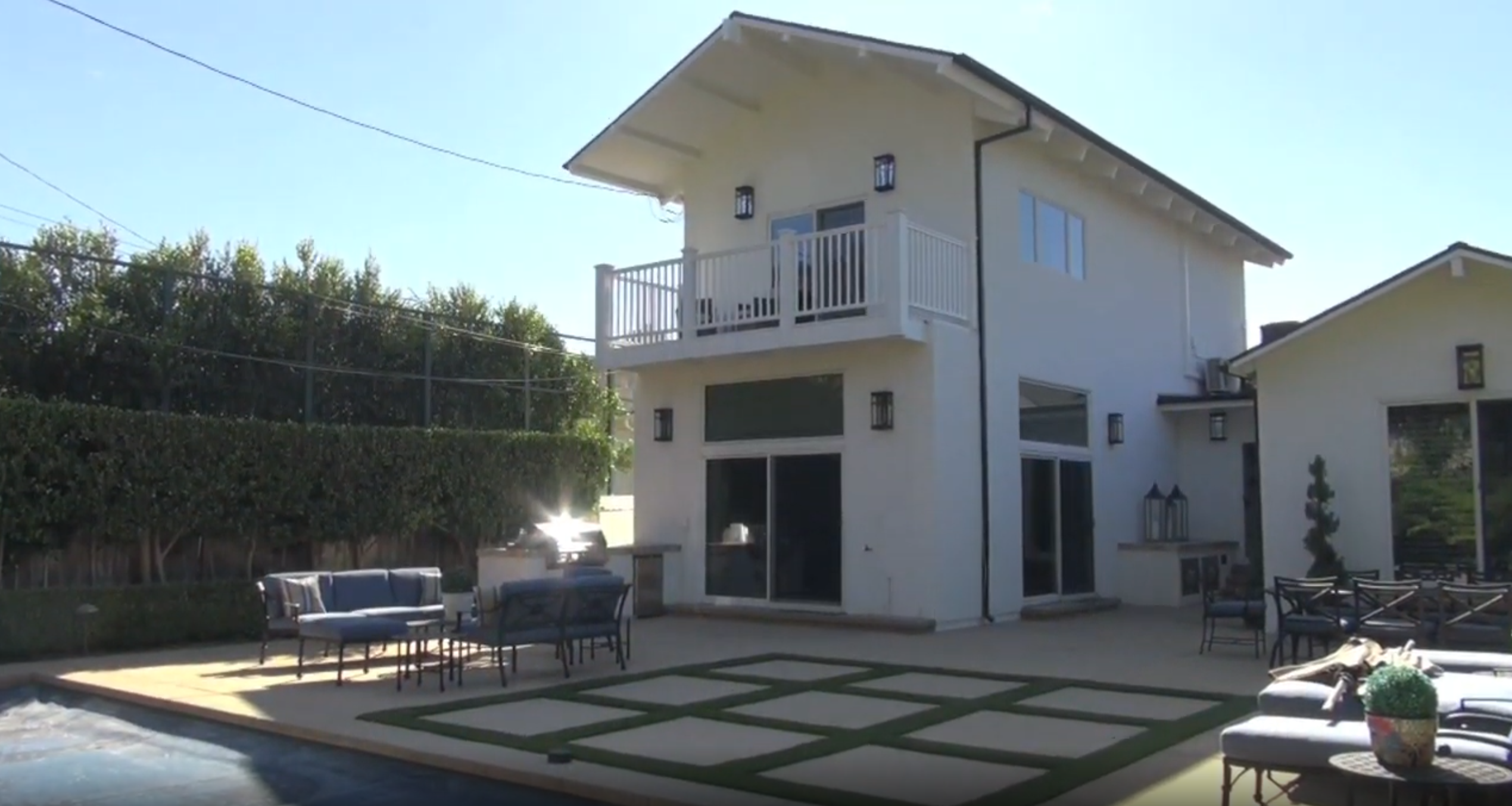 This rear view of the property showcases a two-story residence with a clean, contemporary aesthetic and crisp white siding. The home features a large balcony on the second floor and extensive sliding glass doors on the ground level, ideal for indoor-outdoor entertaining. The expansive backyard includes a swimming pool, a modern paver-and-turf patio area, and comfortable outdoor seating, creating a serene and private oasis.