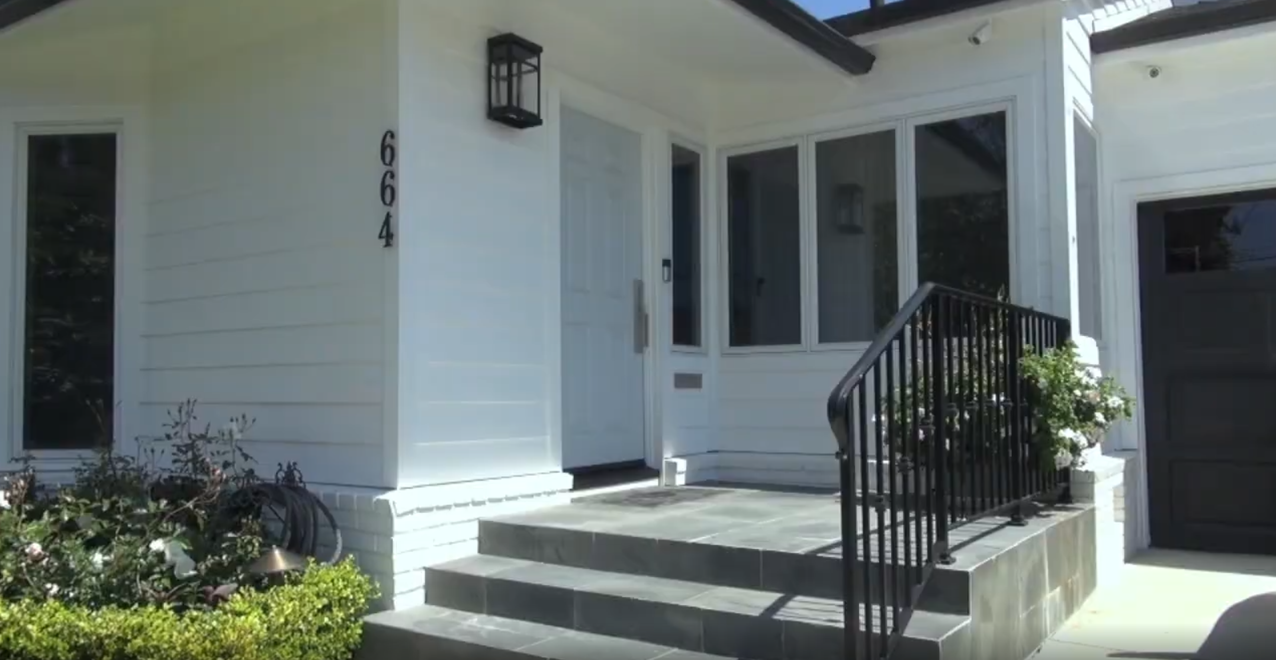 This inviting home exterior features a clean, white-sided facade with a modern light-colored front door and classic house numbers displayed vertically. A set of slate-tiled stairs leads up to the entrance, accented by elegant black iron railings, a matching wall sconce, and well-maintained shrubbery. The overall aesthetic is charming and contemporary with a welcoming curb appeal.