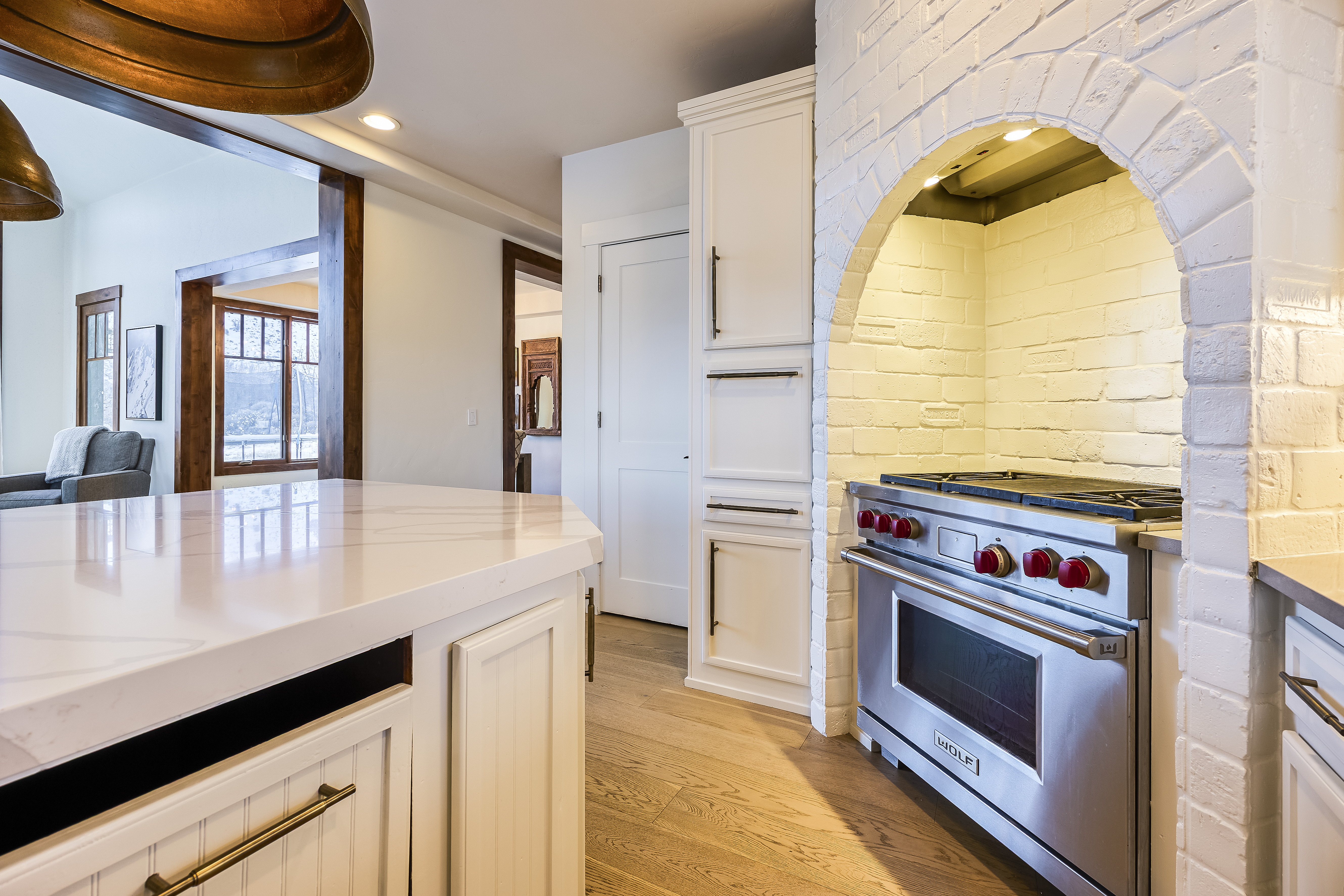 This image showcases a well-appointed kitchen featuring a white brick archway above a professional-grade stove. The kitchen includes custom white cabinetry with dark hardware and light-toned hardwood flooring. A countertop with a light surface is also visible, along with an open doorway leading to another room.