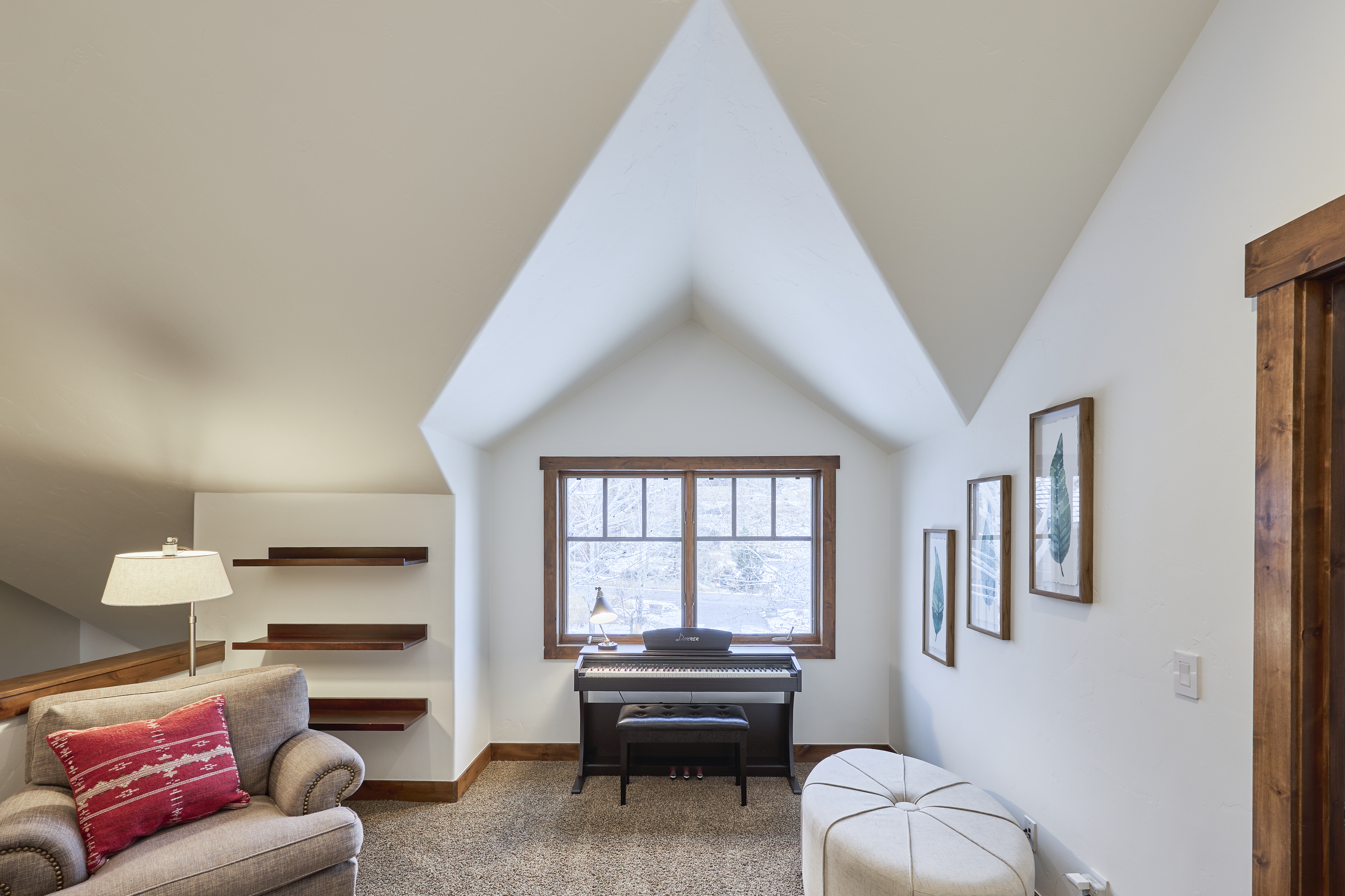 This image showcases a cozy and inviting living room area within a home. A notable feature is the pitched ceiling with a window overlooking a snow-covered landscape, enhancing the room's unique character. Furnishings include a comfortable armchair, a piano, and an ottoman which contribute to the room's relaxed atmosphere.
