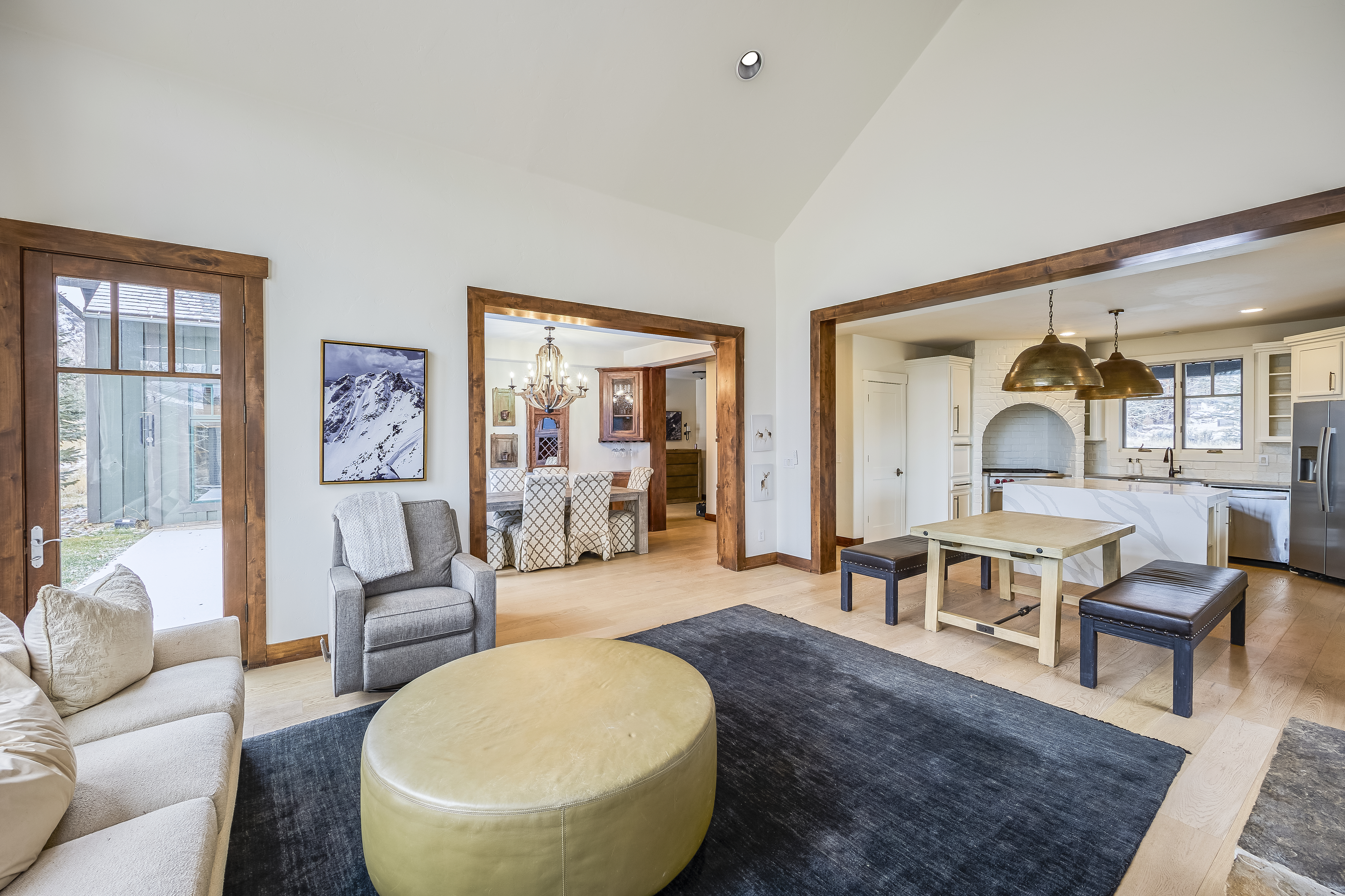 This is a bright and open living area that flows into the dining room and kitchen. The space is well-furnished with a sofa, armchair, ottoman, and a stylish rug. The architectural details include wood-framed doorways and hardwood floors, contributing to a warm and inviting atmosphere.