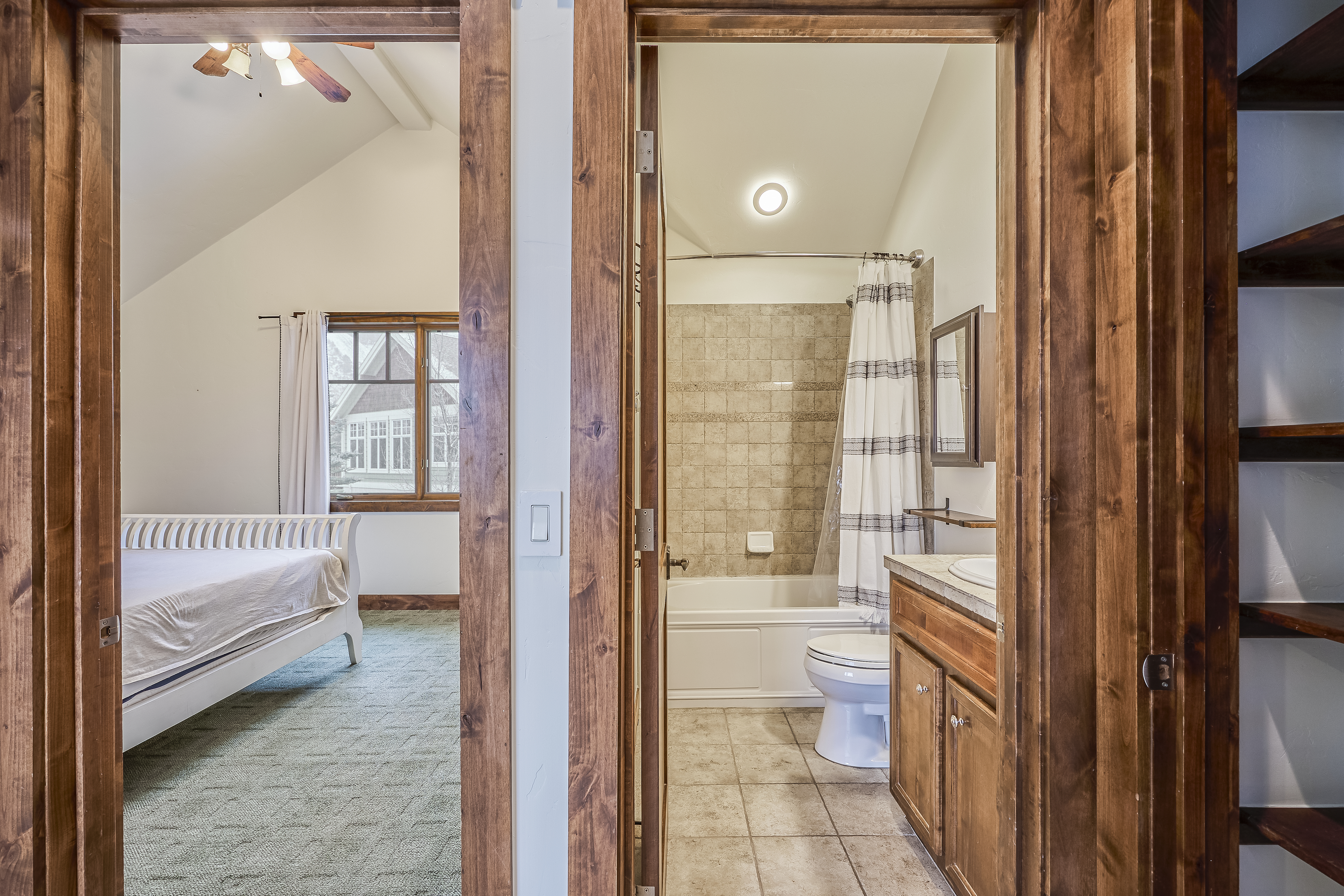 The image showcases a view into a guest bathroom. The bathroom features a bathtub with a shower curtain, a toilet, and a vanity with wooden cabinets and a light-colored countertop. Through an open doorway, a bedroom can be seen, contributing to a sense of connectivity and space.