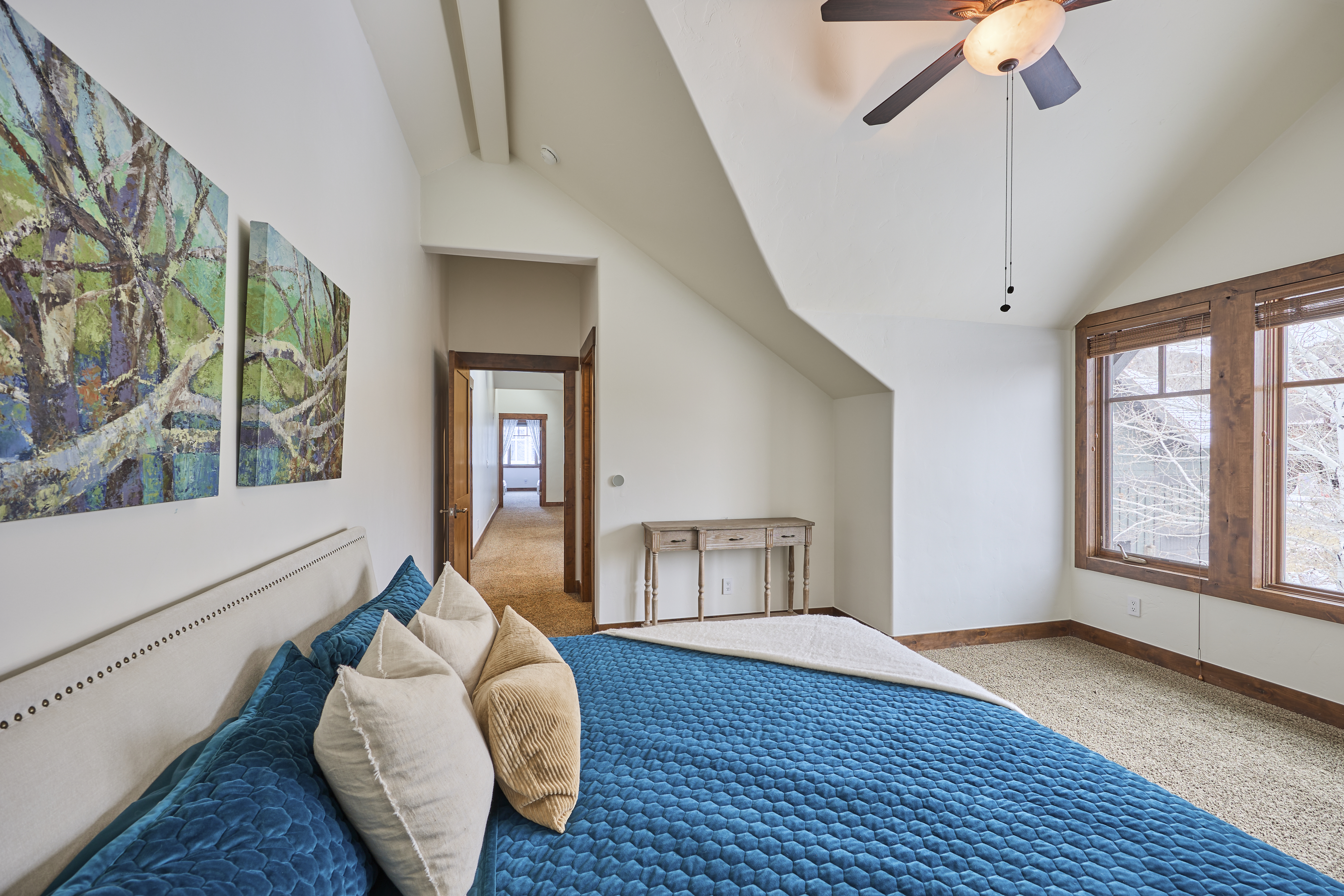 This is a cozy guest bedroom with a blue quilted bedspread and decorative pillows. The room features natural light from a window with wooden trim, abstract artwork on the wall, and a hallway visible through an open doorway. A ceiling fan provides ventilation, and the overall ambiance is comfortable and inviting.