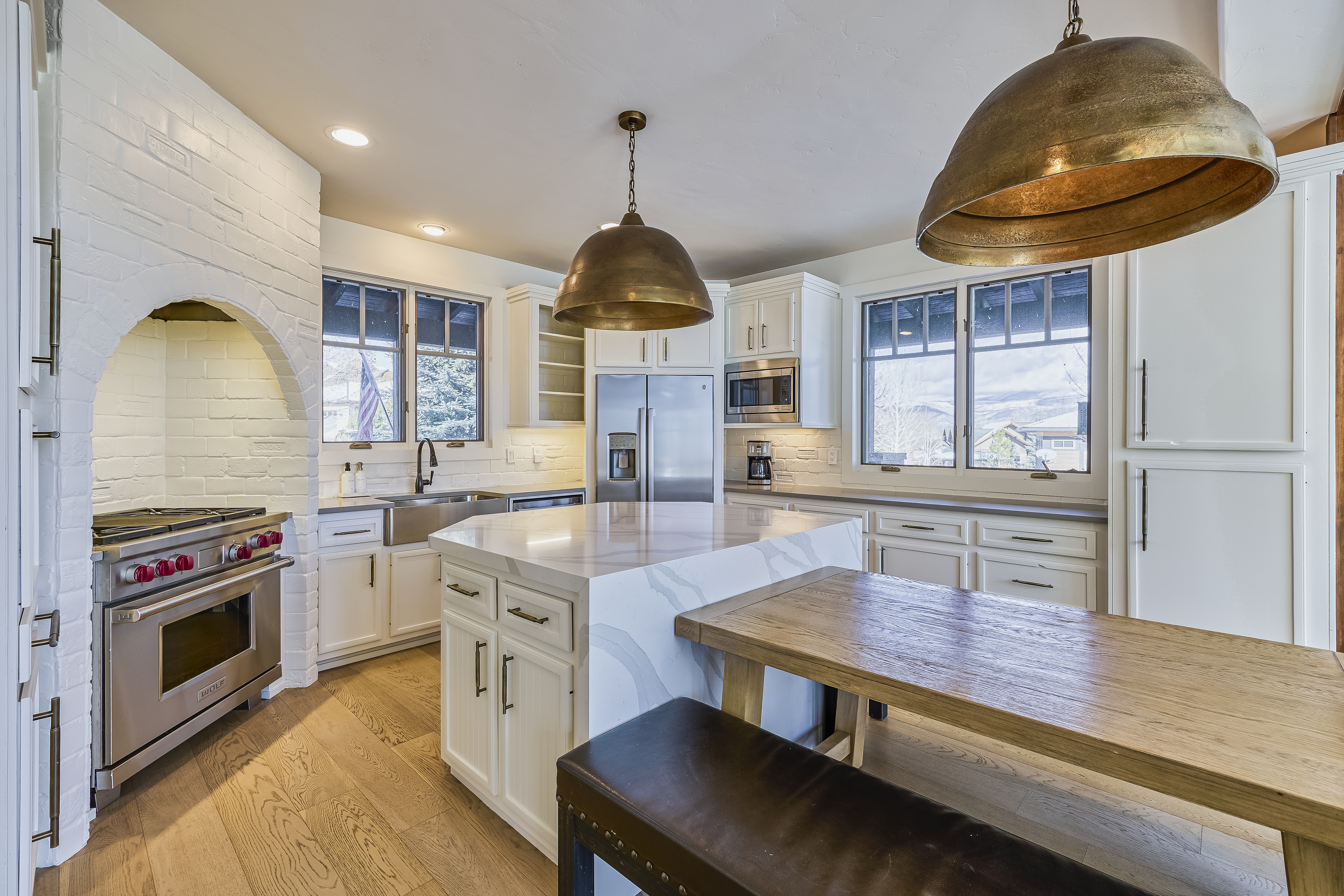 This is a well-lit, modern kitchen featuring white cabinetry, stainless steel appliances, and a contrasting dark countertop. A large kitchen island with a waterfall edge provides ample workspace and seating. The room also includes statement pendant lighting and a natural wood table, creating a warm and inviting atmosphere.