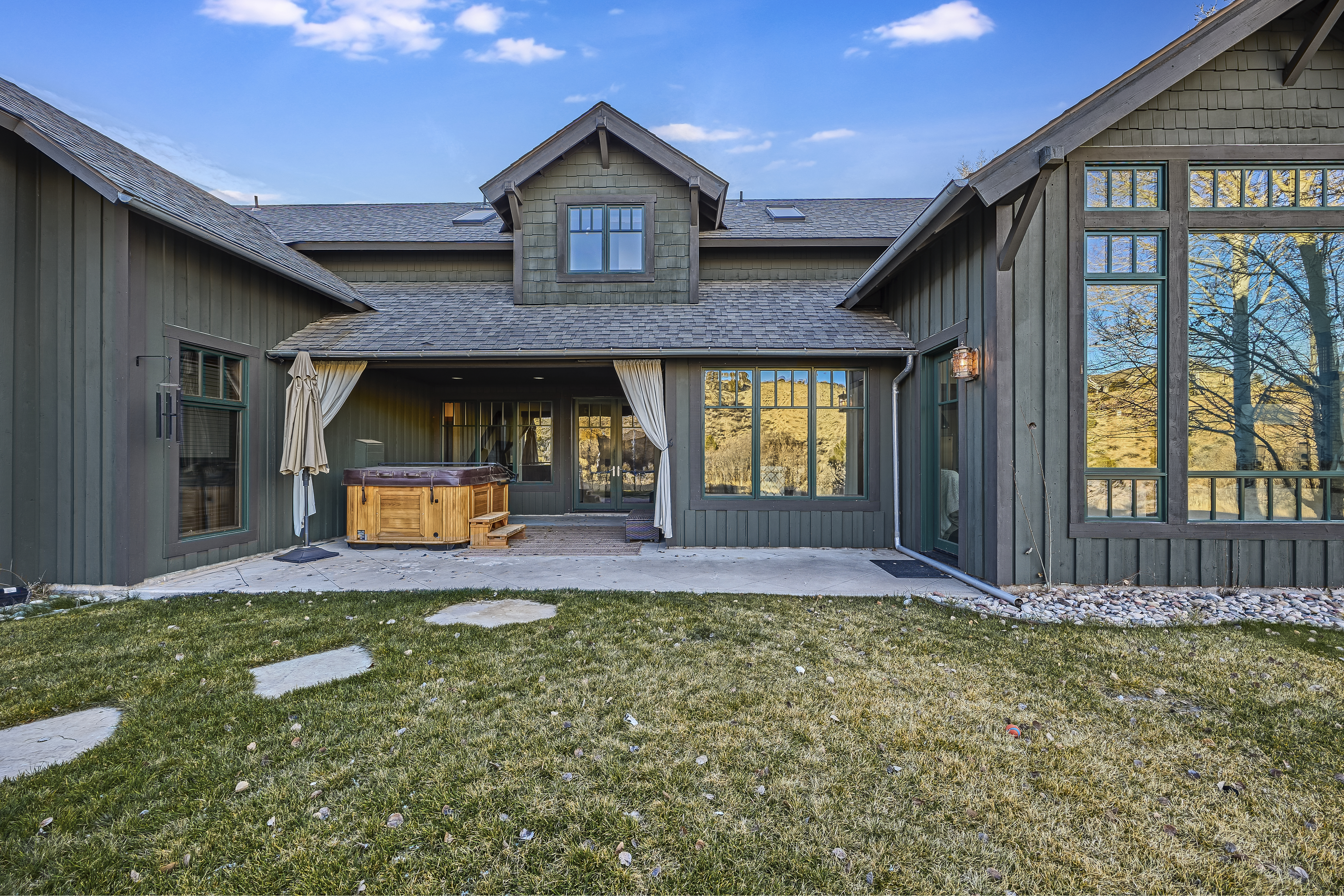 This is a rear view of a home with an outdoor patio and spa. The house features dark green siding, a shingled roof, and large windows that reflect the surrounding landscape. The patio is furnished with a wood-paneled hot tub, and the yard is a mix of grass and stone walkways.
