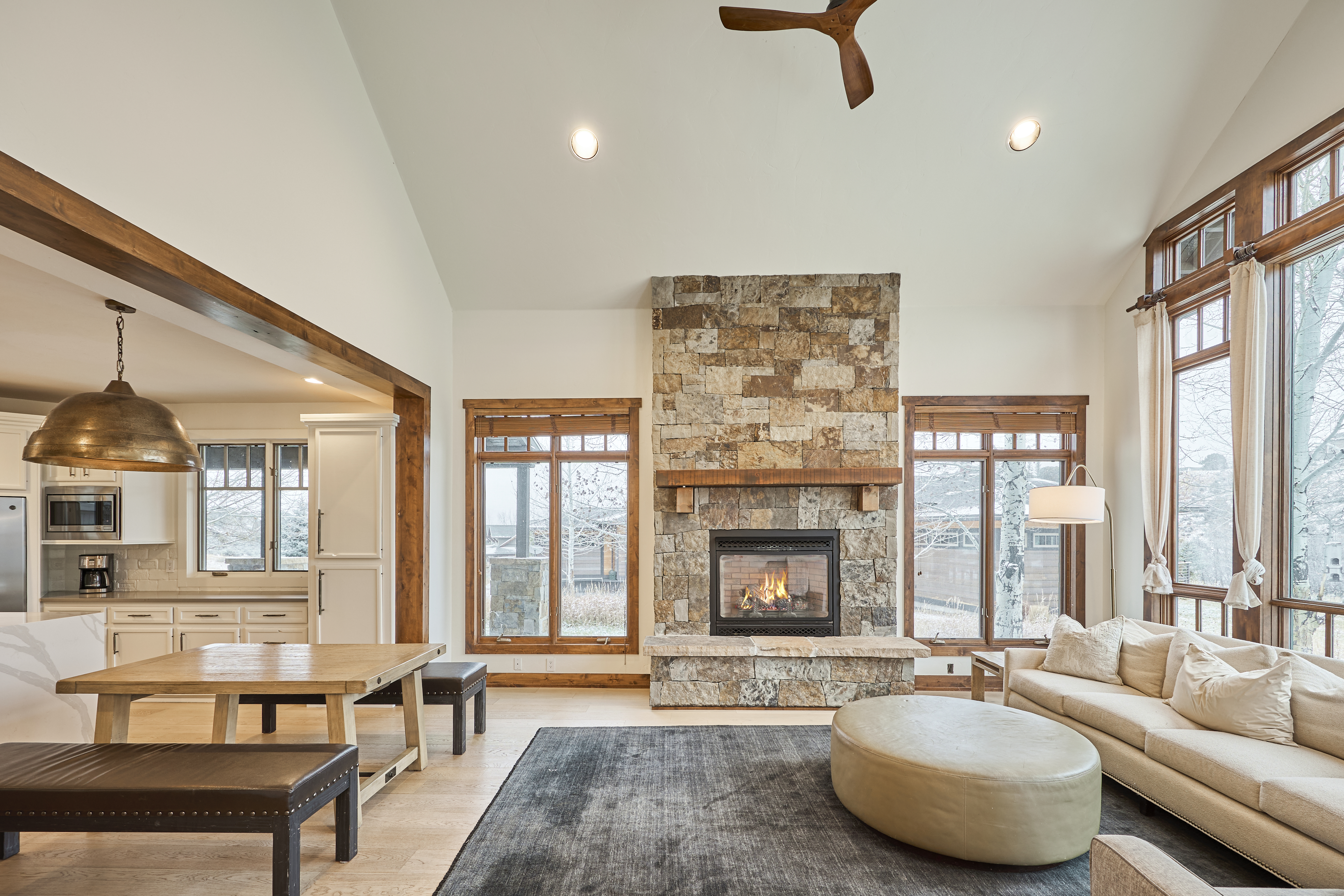 This is a bright and spacious living room featuring a stone fireplace, hardwood floors, and large windows that offer ample natural light. The room is furnished with a beige sofa, an ottoman, a table with benches, and a grey area rug, creating a comfortable and inviting living space. The open layout connects seamlessly to the kitchen, enhancing the home's sense of flow.