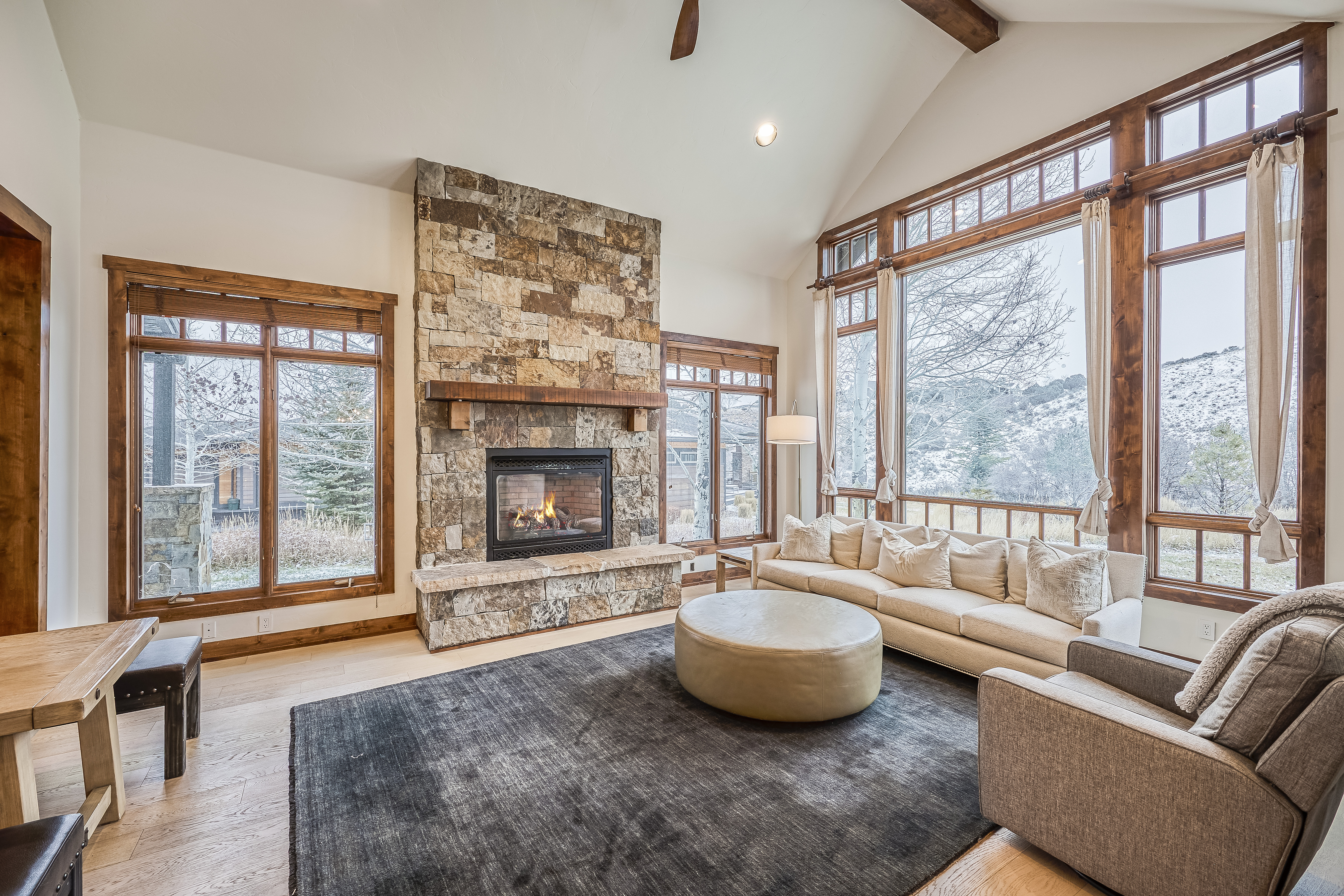 This inviting living room showcases a stone fireplace as a central feature, complemented by large windows that offer natural light and scenic views. The neutral color palette with hardwood floors and a comfortable sofa contributes to a cozy and sophisticated ambiance. The room is well-appointed and perfect for relaxation or entertaining guests.