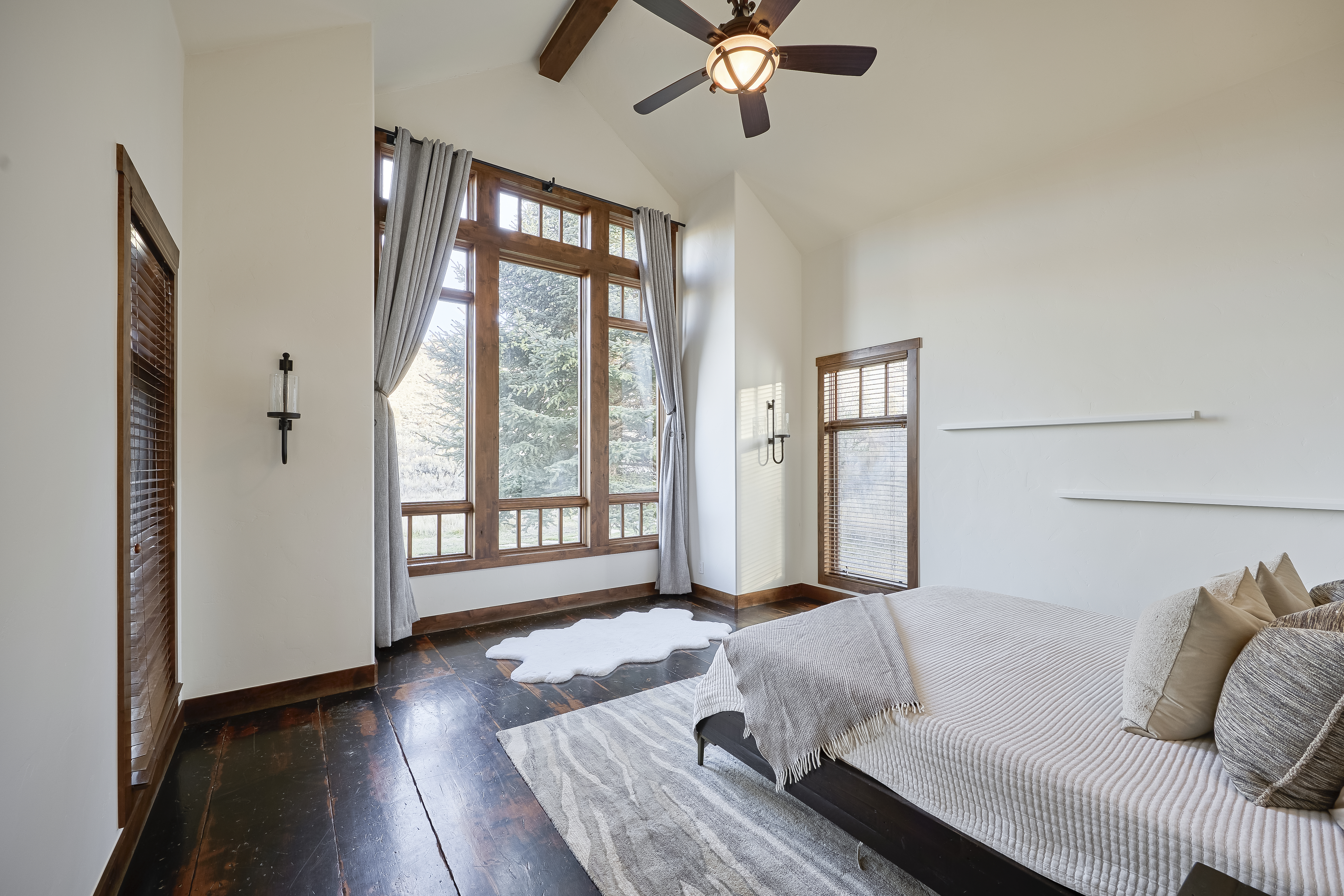 This is a cozy primary bedroom featuring dark wood floors and a vaulted ceiling with exposed beams. A large window offers generous natural light and views presumably of trees or a landscape. The room is furnished with a bed, and the color palette is neutral creating a relaxing atmosphere.