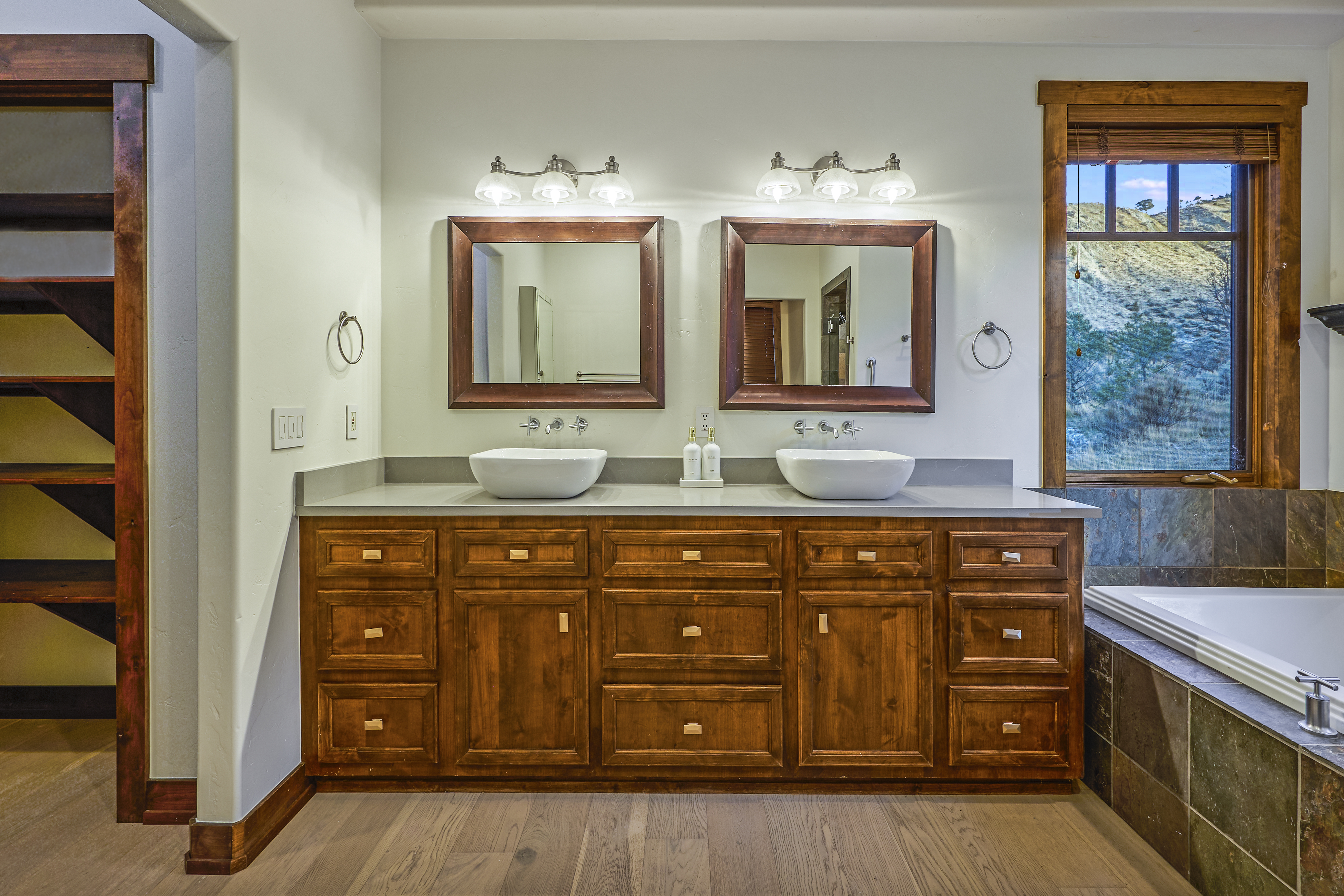 The image presents a well-lit primary bathroom with a double vanity featuring wooden cabinetry and light-colored countertop. Two vessel sinks sit atop the vanity, beneath two square mirrors with matching wood frames. A window with a view to the outdoors is by the bathtub.