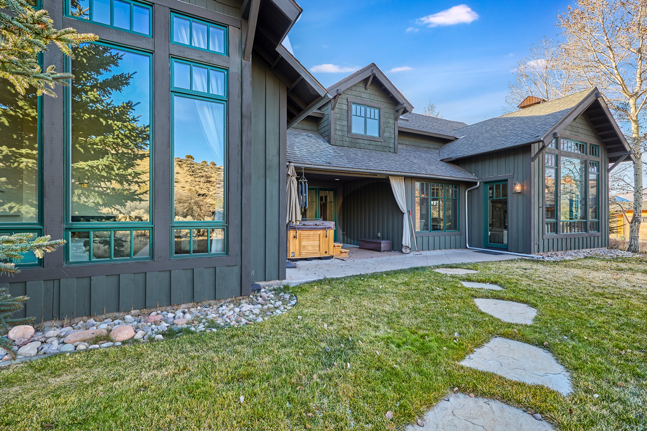 This is a rear exterior view of a well-maintained house, featuring a spacious patio area with a hot tub, stone walkways through the grass, and extensive windows offering views from within. The property showcases a neatly landscaped yard and an inviting outdoor living space perfect for relaxation and entertaining. The house exhibits a craftsman style with muted green horizontal siding and a varied roofline.