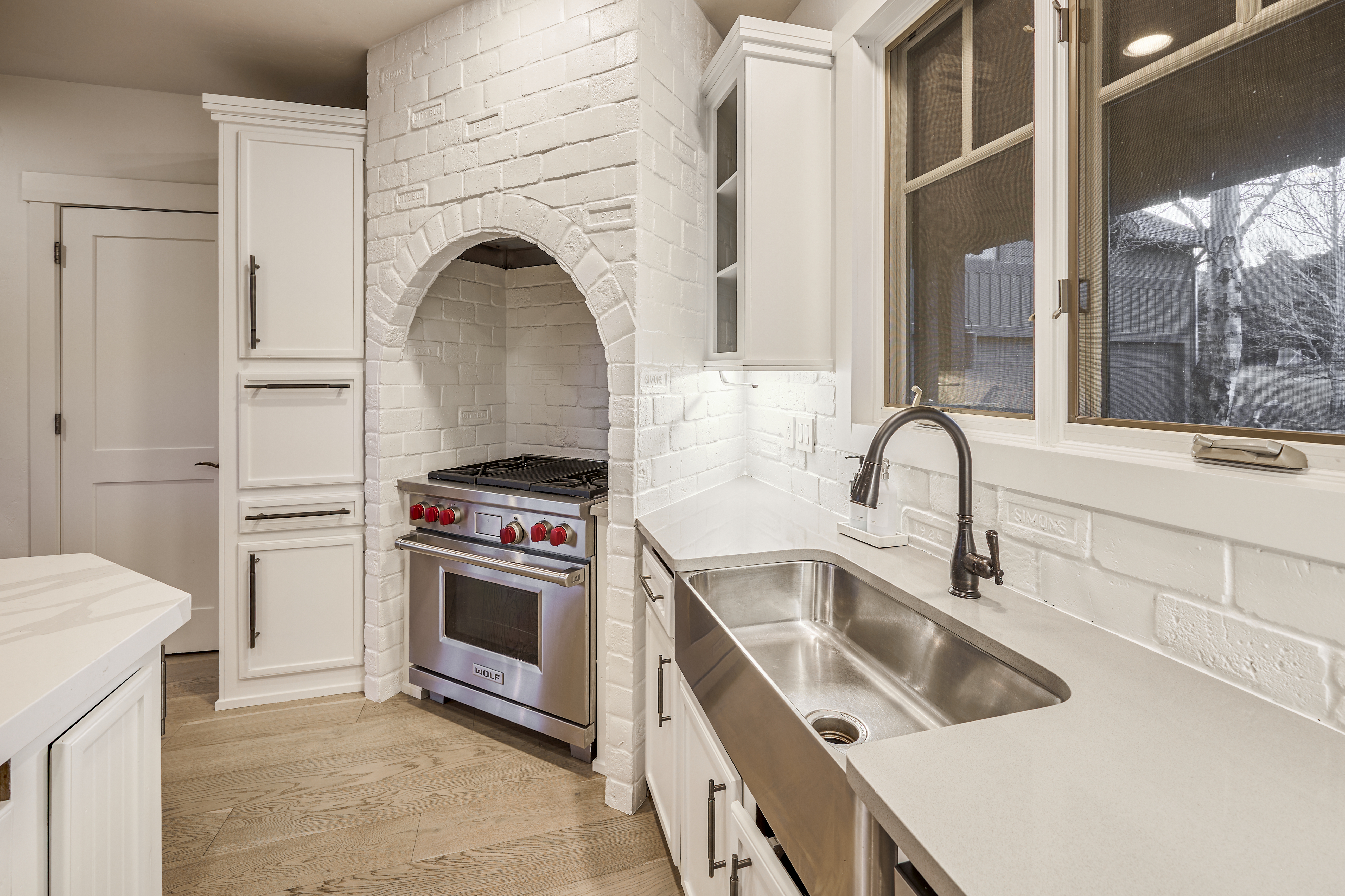 The image showcases a well-lit kitchen featuring white cabinetry and a unique brick-covered alcove with a stainless steel Wolf range. A farmhouse sink with a dark metal faucet is placed beneath a window, adding to the kitchen's charm. The light wood flooring completes the bright and clean aesthetic of the space.