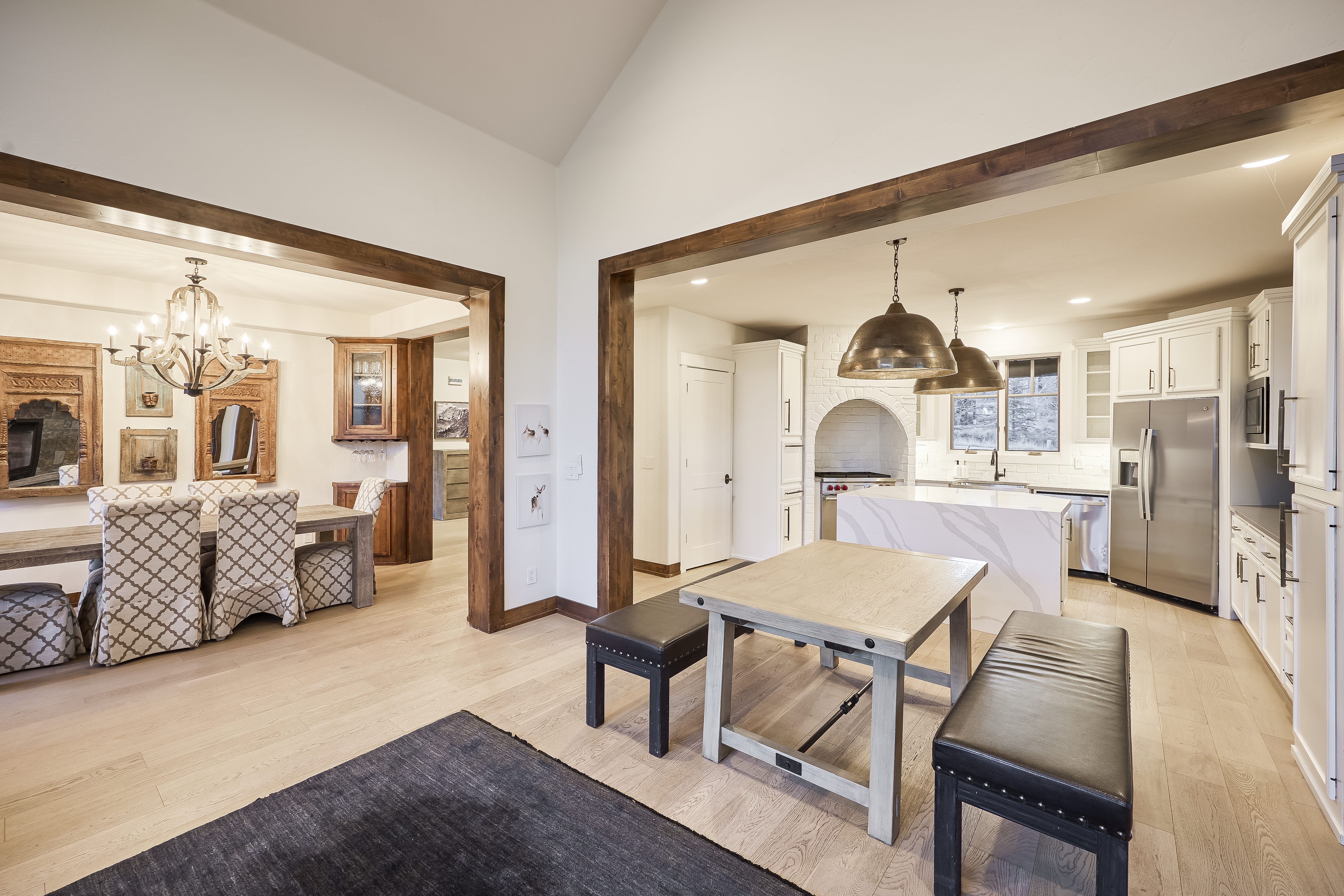 This interior shot showcases a well-lit kitchen and dining area. The kitchen features white cabinetry, stainless steel appliances, and a central island with a unique countertop design. An adjacent dining area includes a wooden table surrounded by upholstered chairs under a chandelier, contributing to a warm and inviting atmosphere.