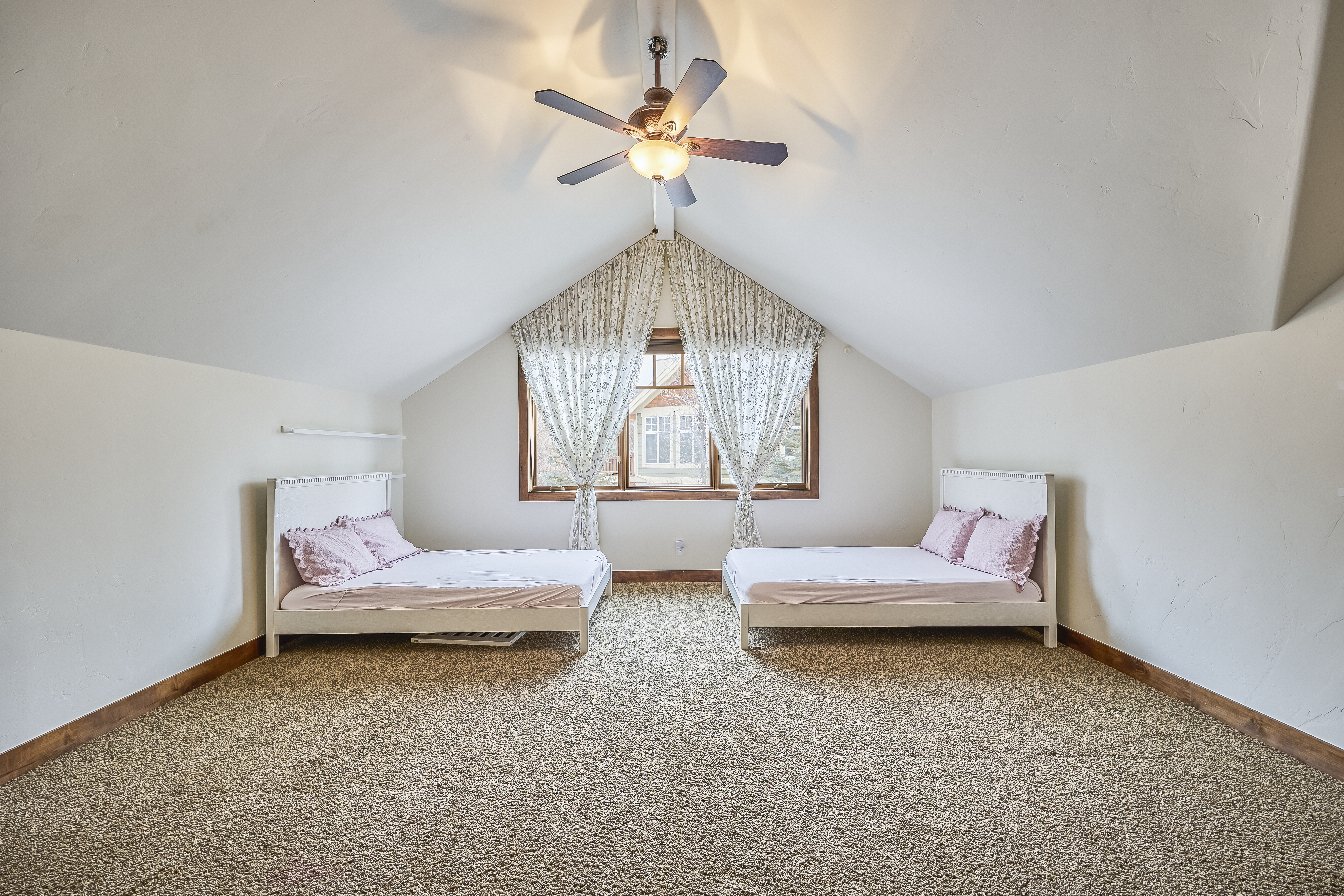 This interior shot showcases a charming guest bedroom within an attic space. The room features two twin-sized beds with white frames, dressed in pink bedding, and is bathed in natural light from a central window adorned with floral curtains. The ceiling fan adds to the room's appeal, while the neutral-toned carpet creates a warm and inviting atmosphere.