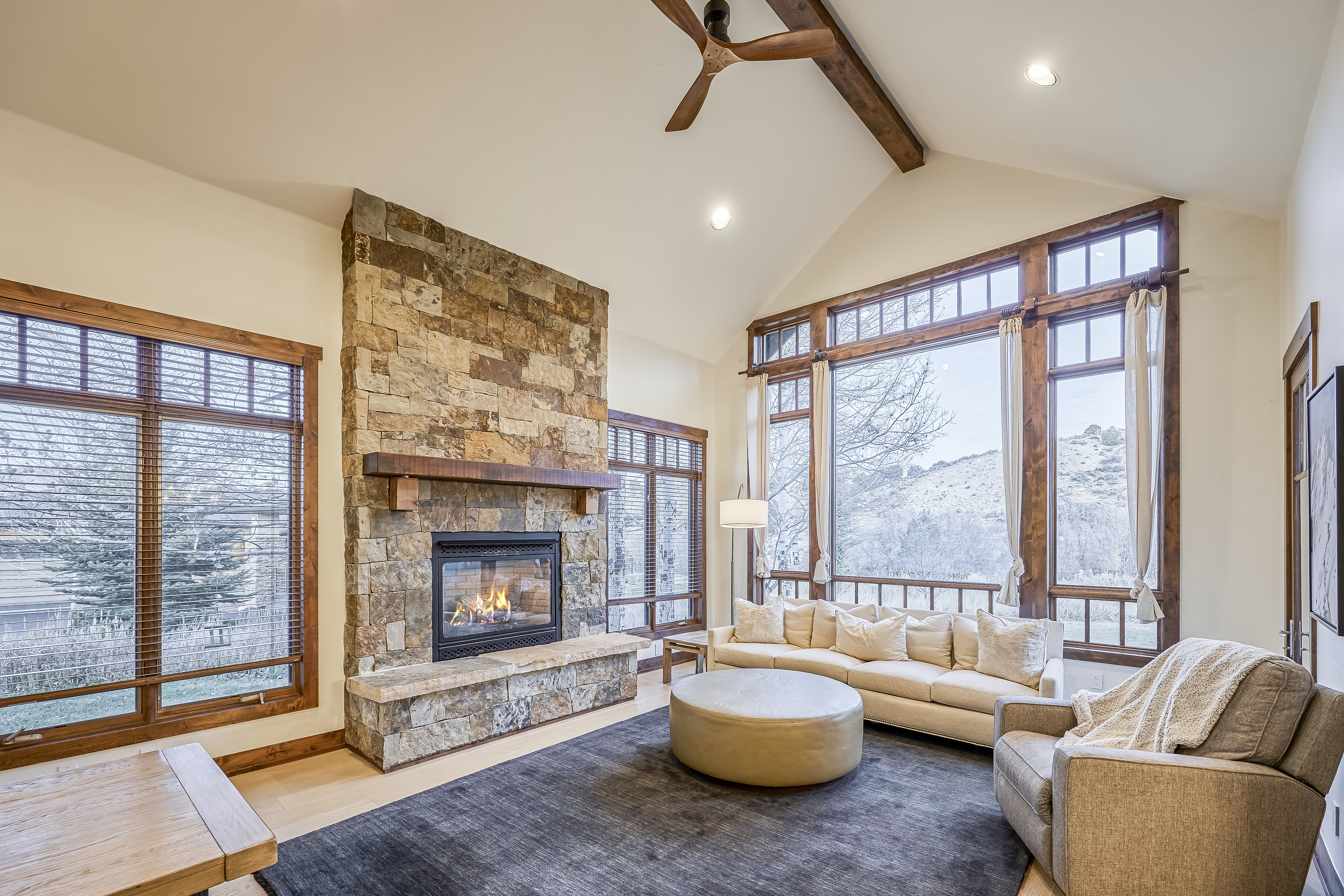 This is a cozy living room with a stone fireplace as its focal point. Large windows provide ample natural light, and the room is furnished with a comfortable-looking sofa, armchair, and ottoman. The decor features natural wood elements, contributing to a warm and inviting atmosphere.