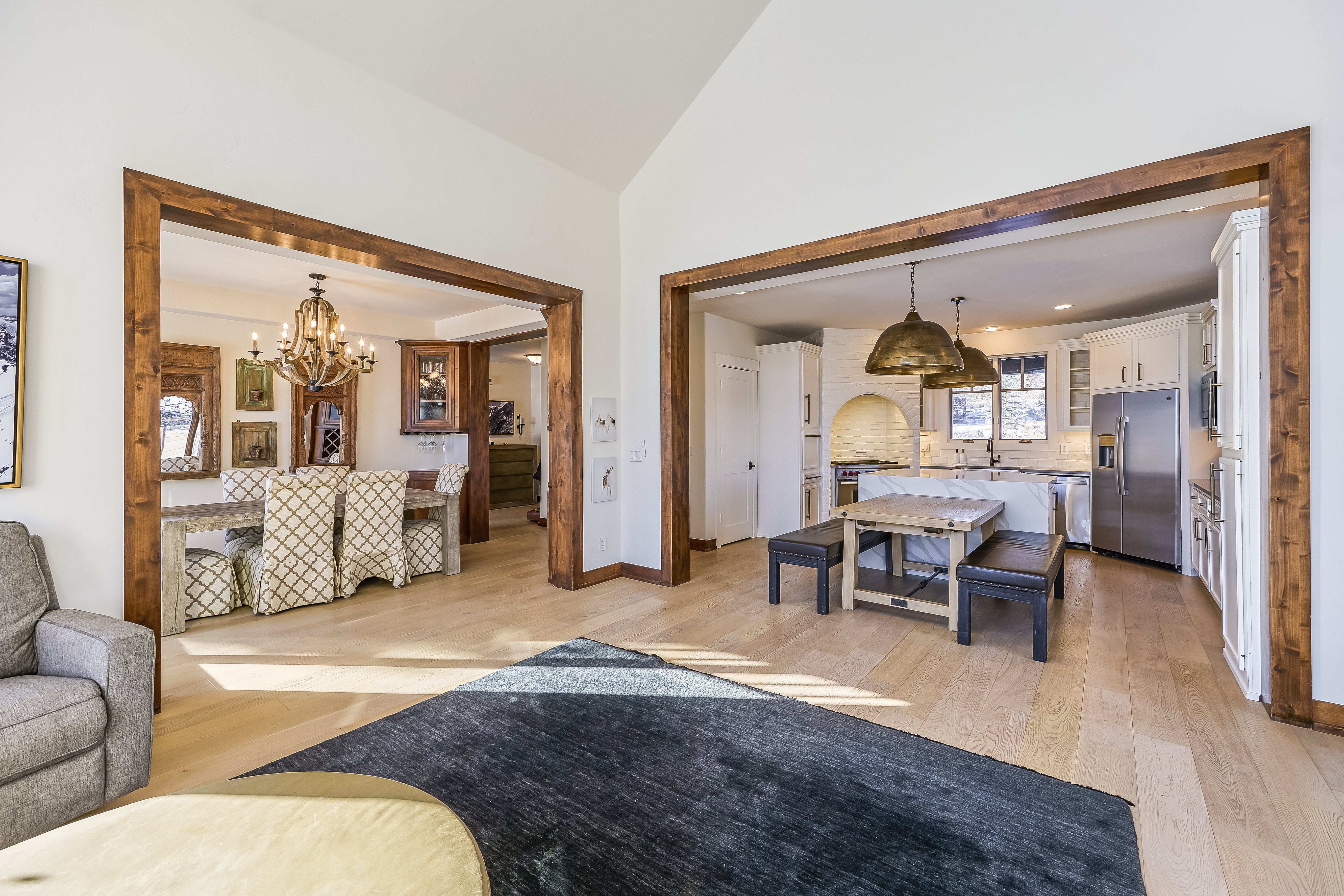 This interior shot showcases a living room that seamlessly connects to the dining and kitchen areas. The open-concept design is accentuated by wooden beams that frame each space, creating a warm and inviting atmosphere. Light hardwood floors and neutral wall colors provide a bright backdrop for stylish furniture and decor, perfect for family living and entertaining.