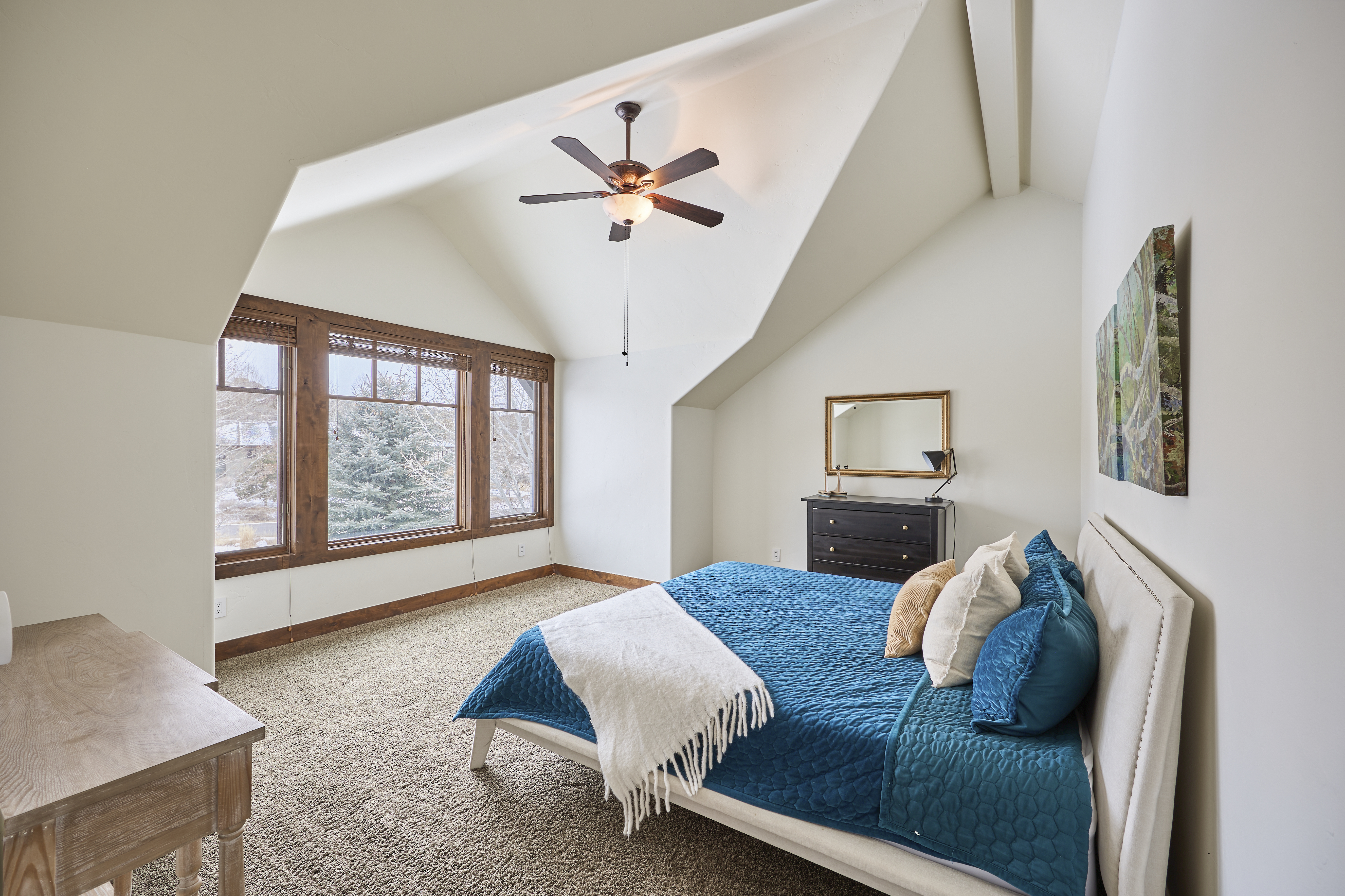 This is a well-lit primary bedroom featuring a bed with a blue patterned cover and several pillows. The room includes a dark wood dresser with a mirror above it and artwork on the wall. Windows with dark wood frames let in natural light, and the walls are painted a neutral color, creating a calming and inviting atmosphere.