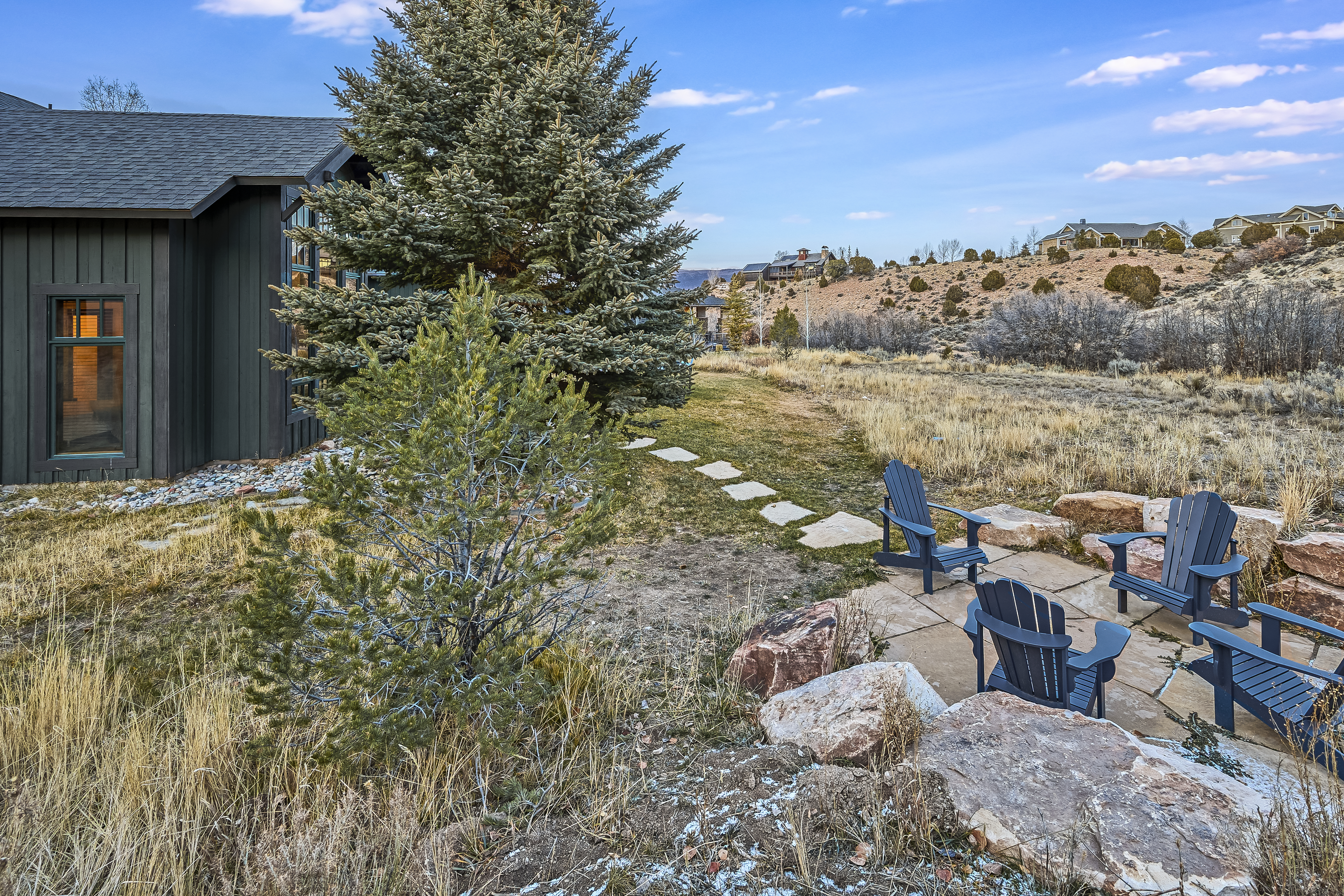 The image showcases the rear exterior of a home with a well-manicured yard. Adirondack chairs are arranged around a paved patio, suggesting an inviting outdoor living space. Mature trees provide privacy and enhance the property's appeal, creating a serene and relaxing atmosphere.