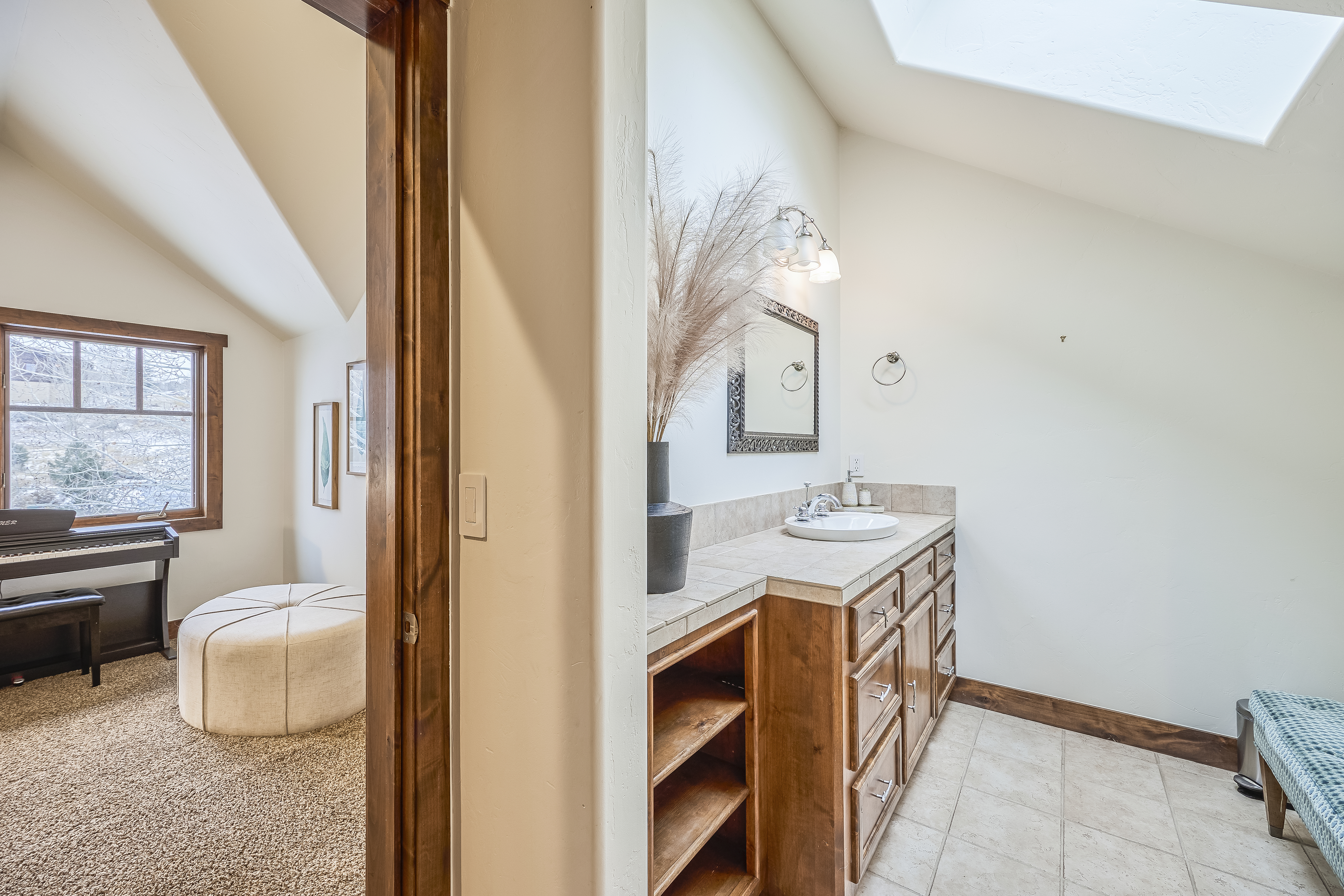 The image showcases a well-lit guest bathroom with a vanity featuring a countertop sink, wood cabinetry, and open shelving. Natural light streams in from a skylight. A glimpse into an adjacent room reveals a piano and upholstered ottoman, adding to the bright and inviting atmosphere.