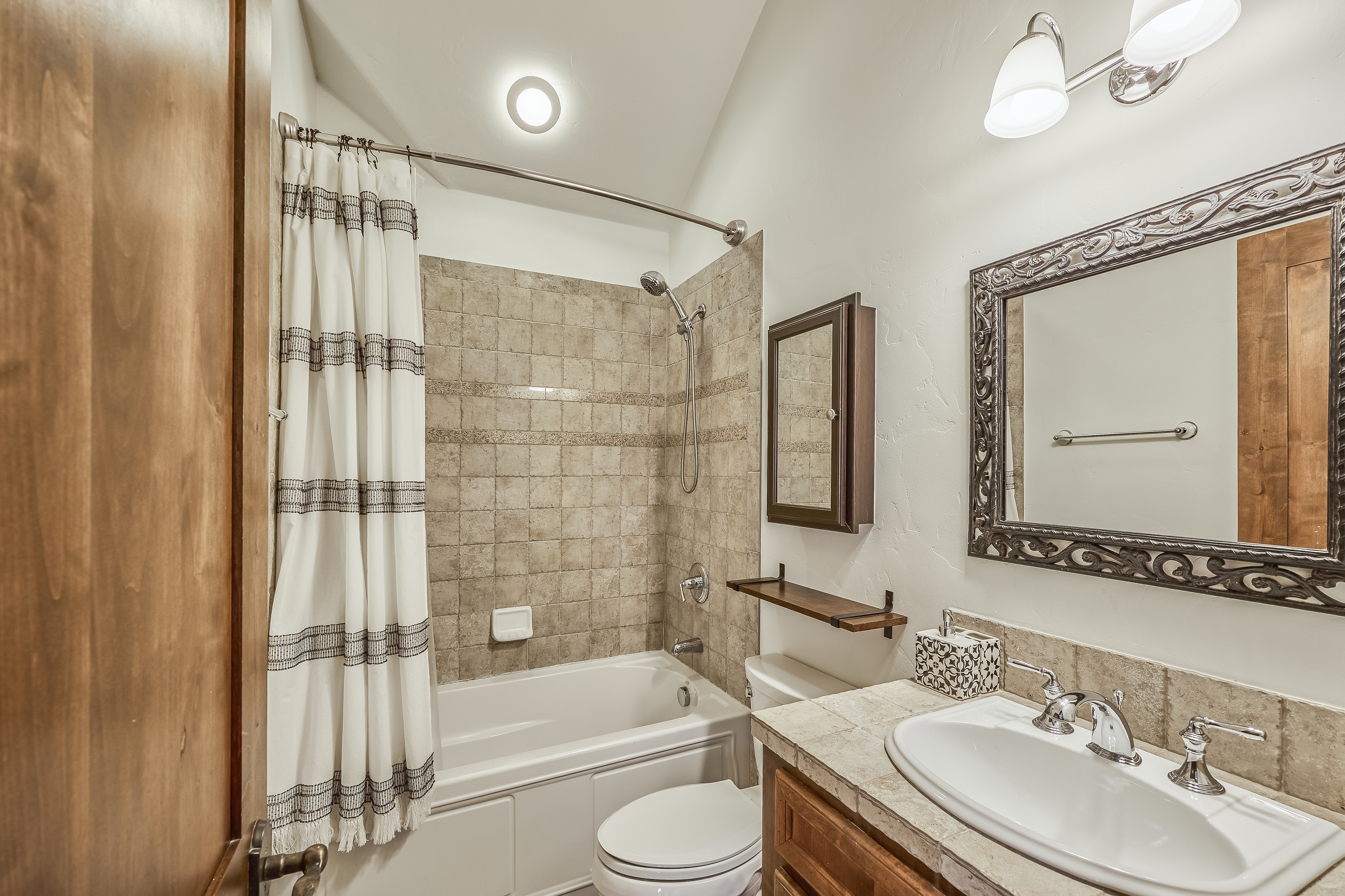 This is a bathroom featuring a tile-surround bathtub with a shower, along with a wooden vanity and a framed mirror. The warm-toned tile imparts a cozy feel, while the ample lighting enhances the space. A toilet and a small storage shelf next to it are also visible.