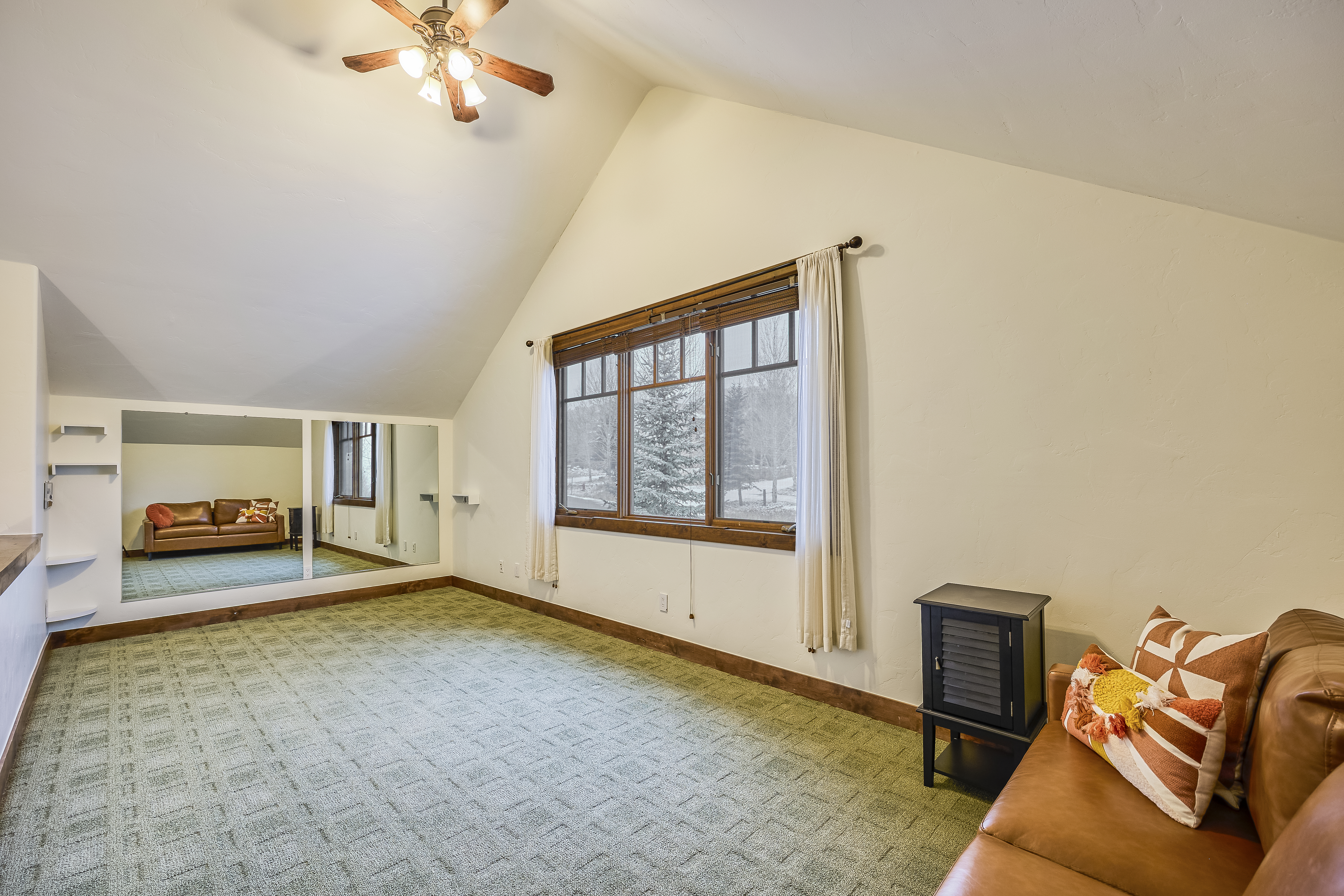 This interior shot showcases a guest bedroom or other bedroom. The room features a green patterned carpet, a window with neutral drapes, and a mirrored wall adding to the sense of space. The high, angled ceiling and ceiling fan are notable architectural elements.