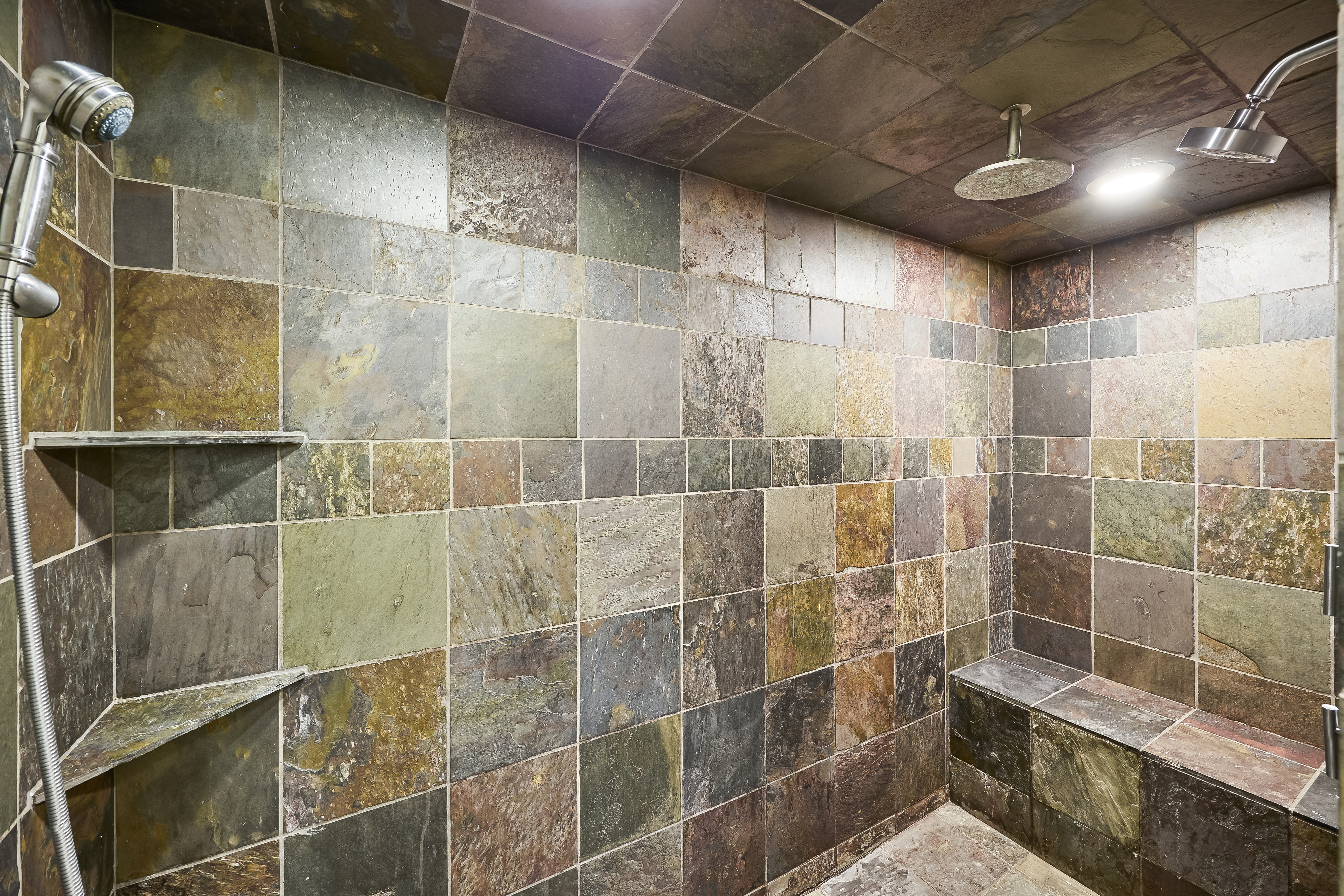 This is a shower room with a tiled stone wall and floor. The shower has a rainfall-style head, with an additional sprayer. There are two shelves along the left wall, as well as a built-in bench on the right side.