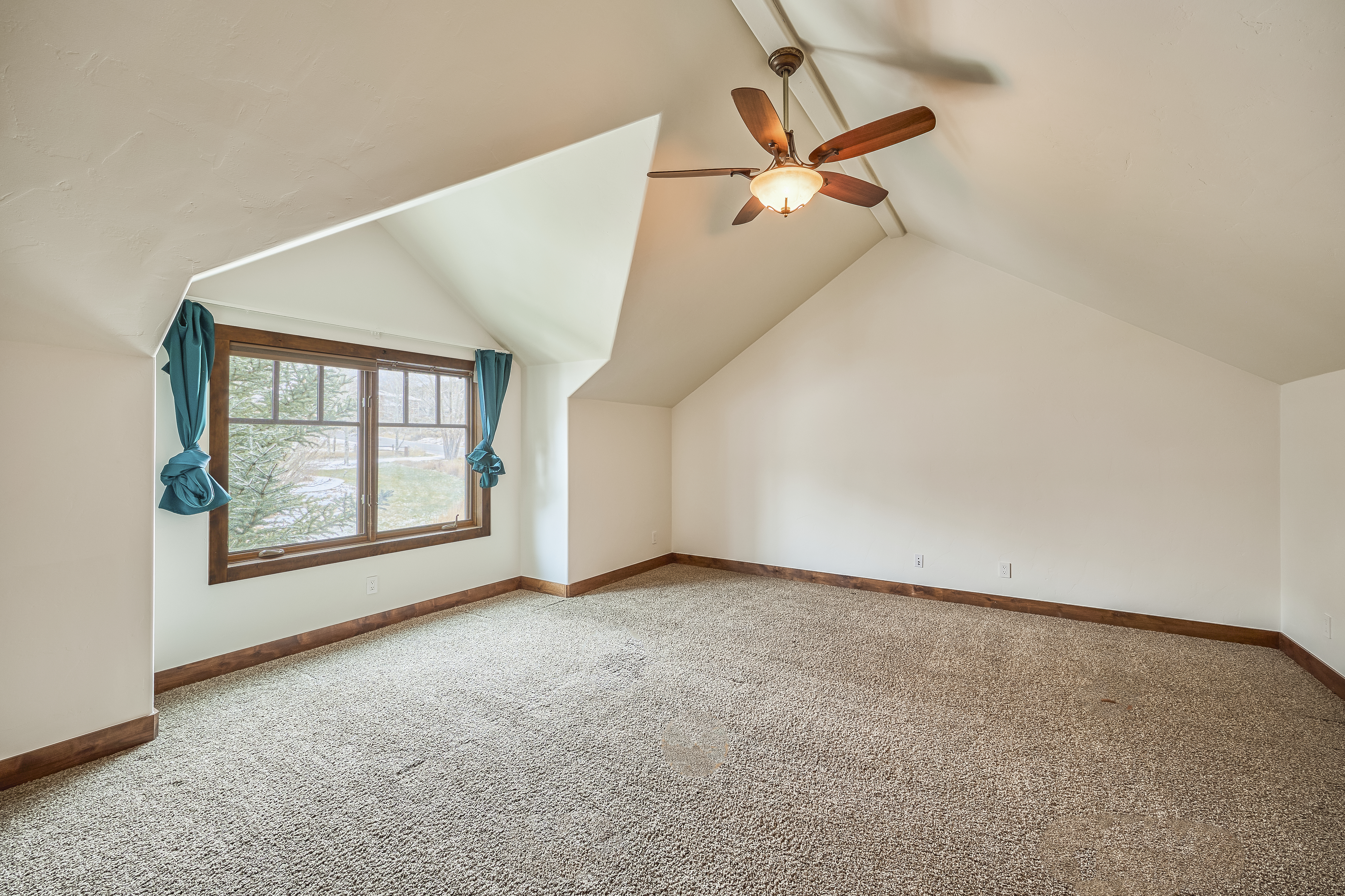 This is a bright and airy bedroom featuring an angled ceiling, a window with blue curtains and brown trim. The brown trim is used along the base of the walls as well. The room has neutral paint and carpet, and there is a ceiling fan in the center of the ceiling.