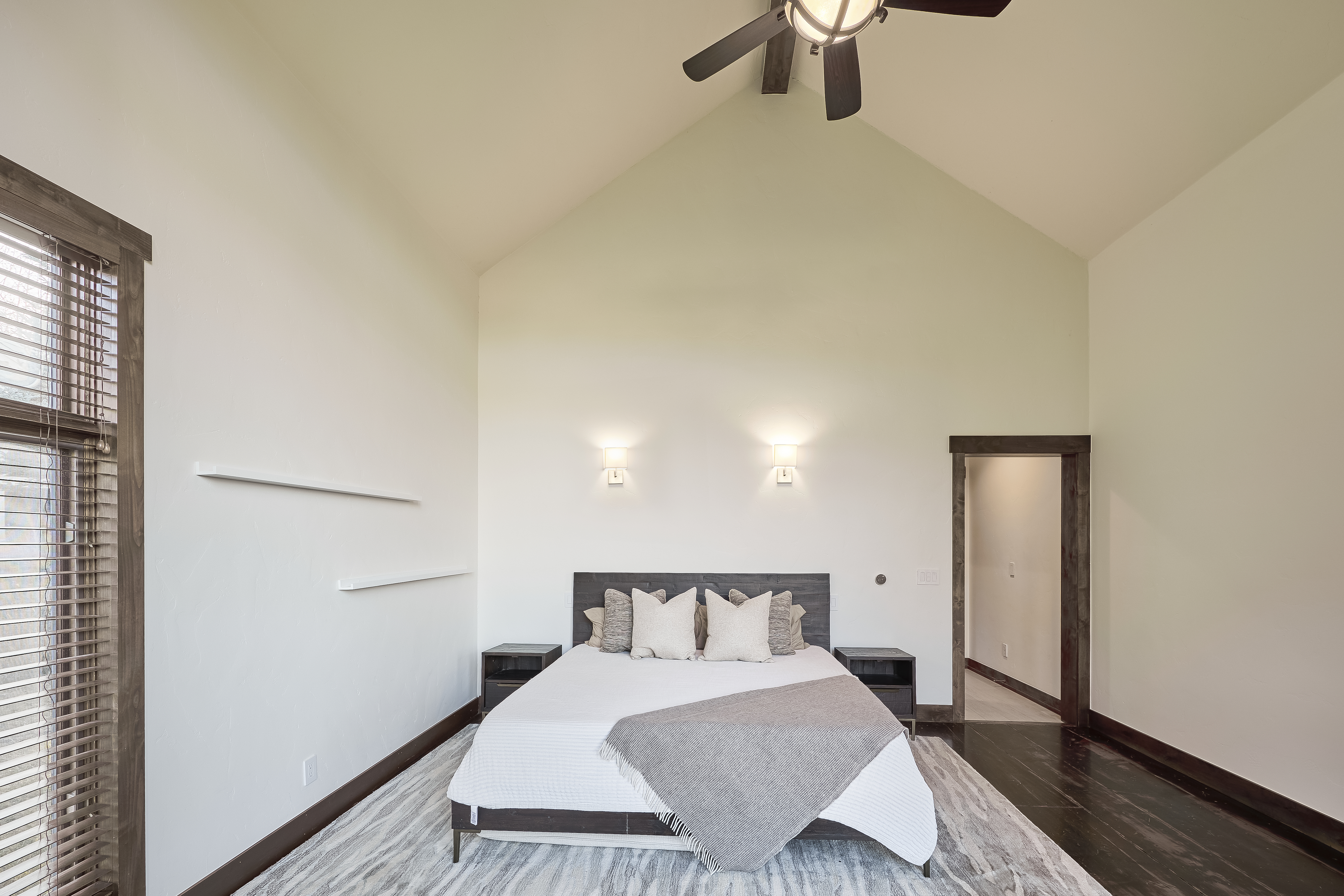 This is a view of a primary bedroom with a high vaulted ceiling. The room features a bed with neutral-toned bedding, two nightstands, and accent lighting sconces. There is a window with blinds and an open door frame, and the room has a modern yet cozy feel.