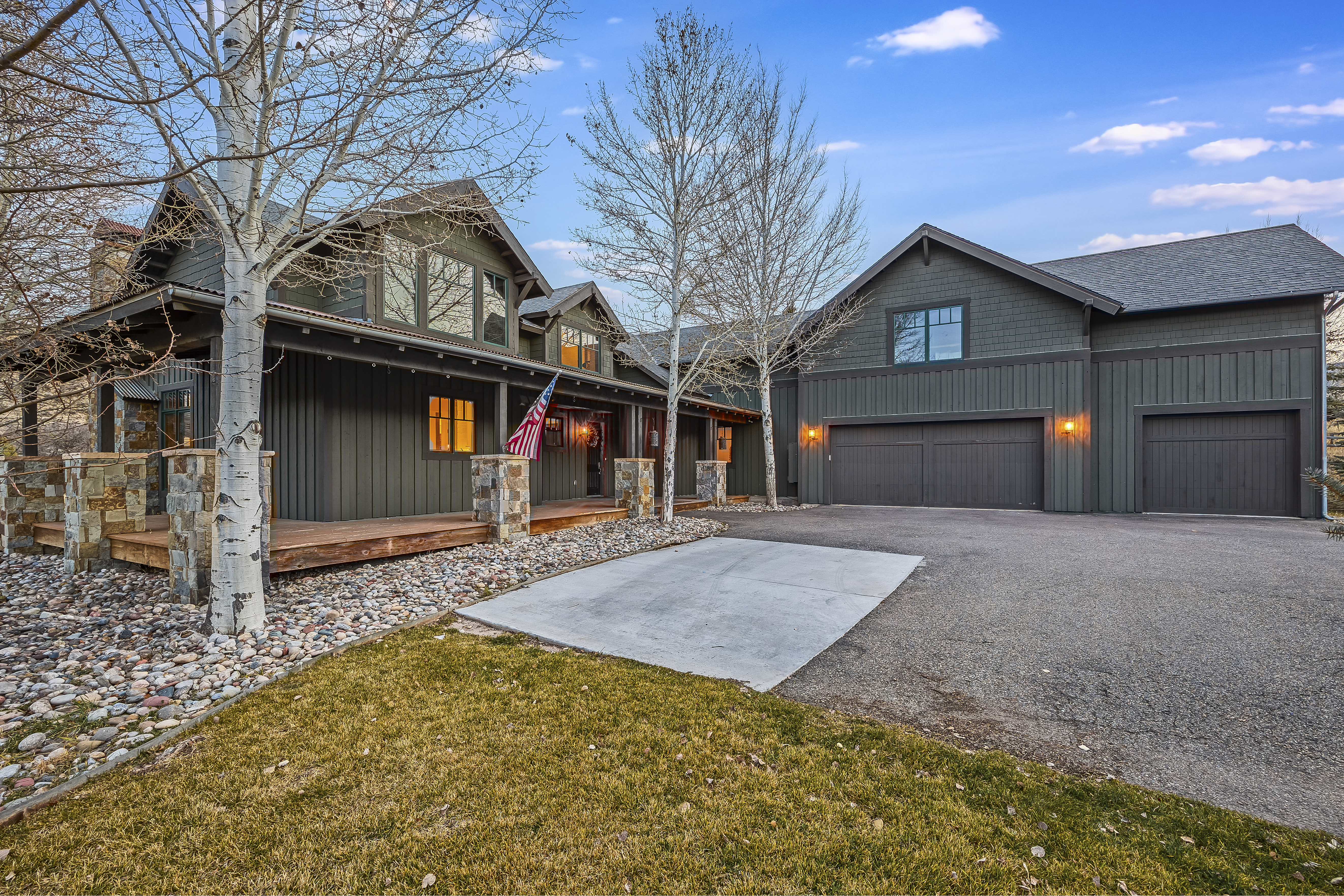 The image showcases the front exterior of a home with a distinctive architectural style. It features a dark-colored facade, stone accents on the porch columns, and a spacious driveway leading to a multi-car garage. Landscaping includes a mix of grass, rocks, and deciduous trees, adding to the property's curb appeal under a clear sky.