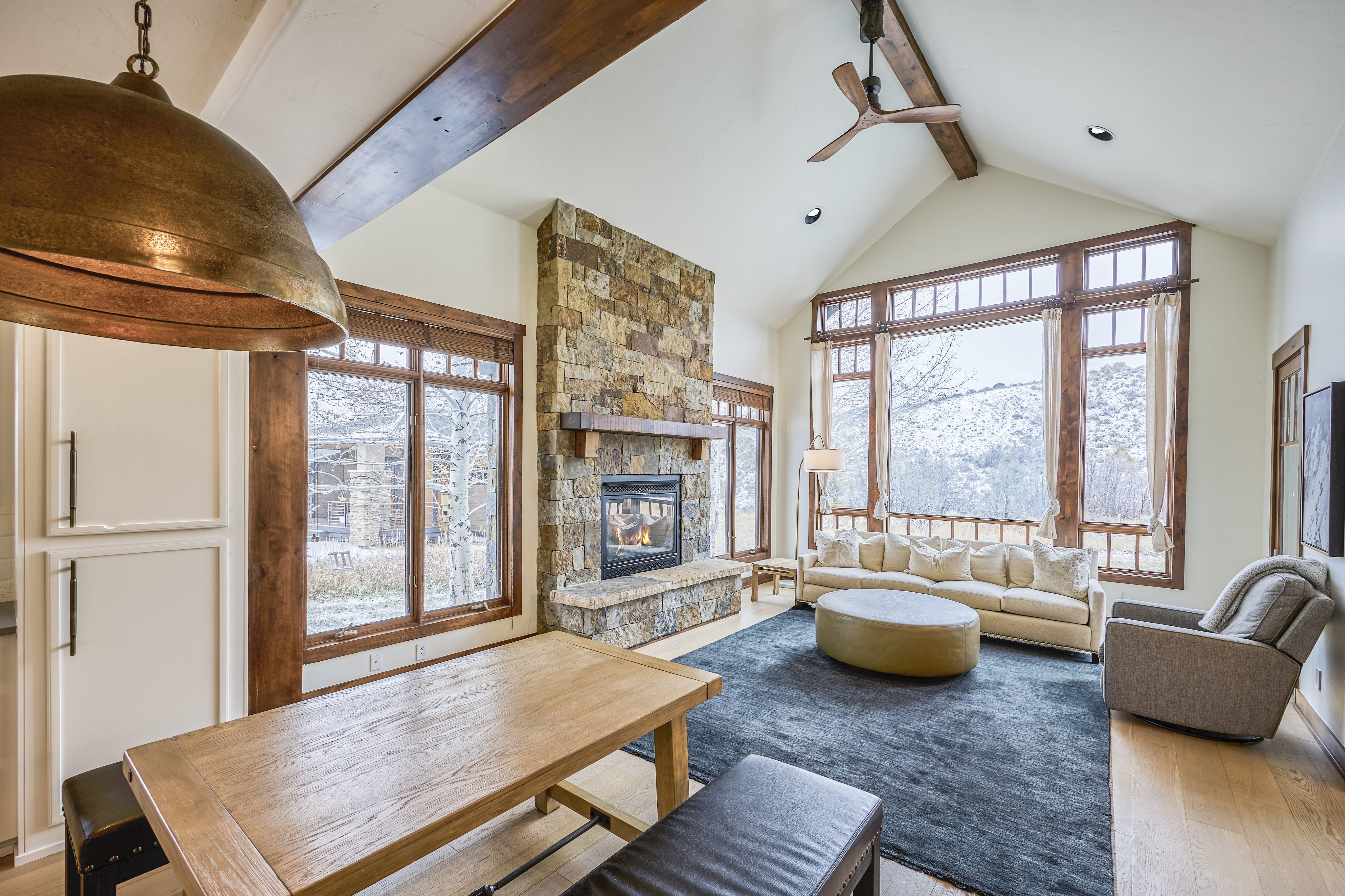 This inviting living room features a stone fireplace, hardwood floors, and large windows offering natural light and views. The room includes comfortable seating around a central ottoman, creating a cozy and upscale atmosphere. Natural wood accents, such as the window trim and ceiling beams, add warmth and character.
