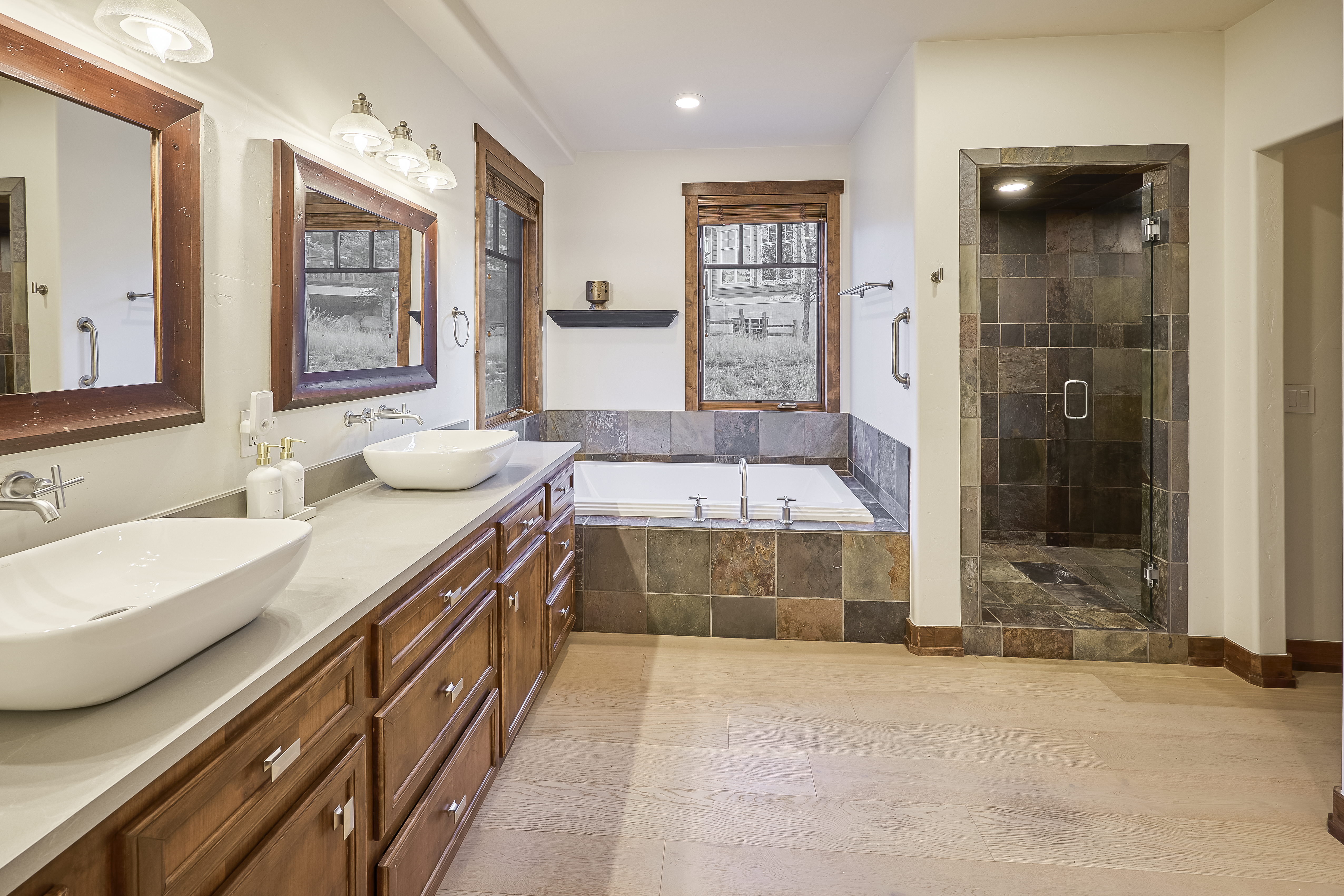 This is a primary bathroom featuring a double vanity with white vessel sinks and wooden cabinets. A soaking tub is surrounded by decorative tile. The adjacent walk-in shower has similar tile and a glass door. The floor is made of light wood.