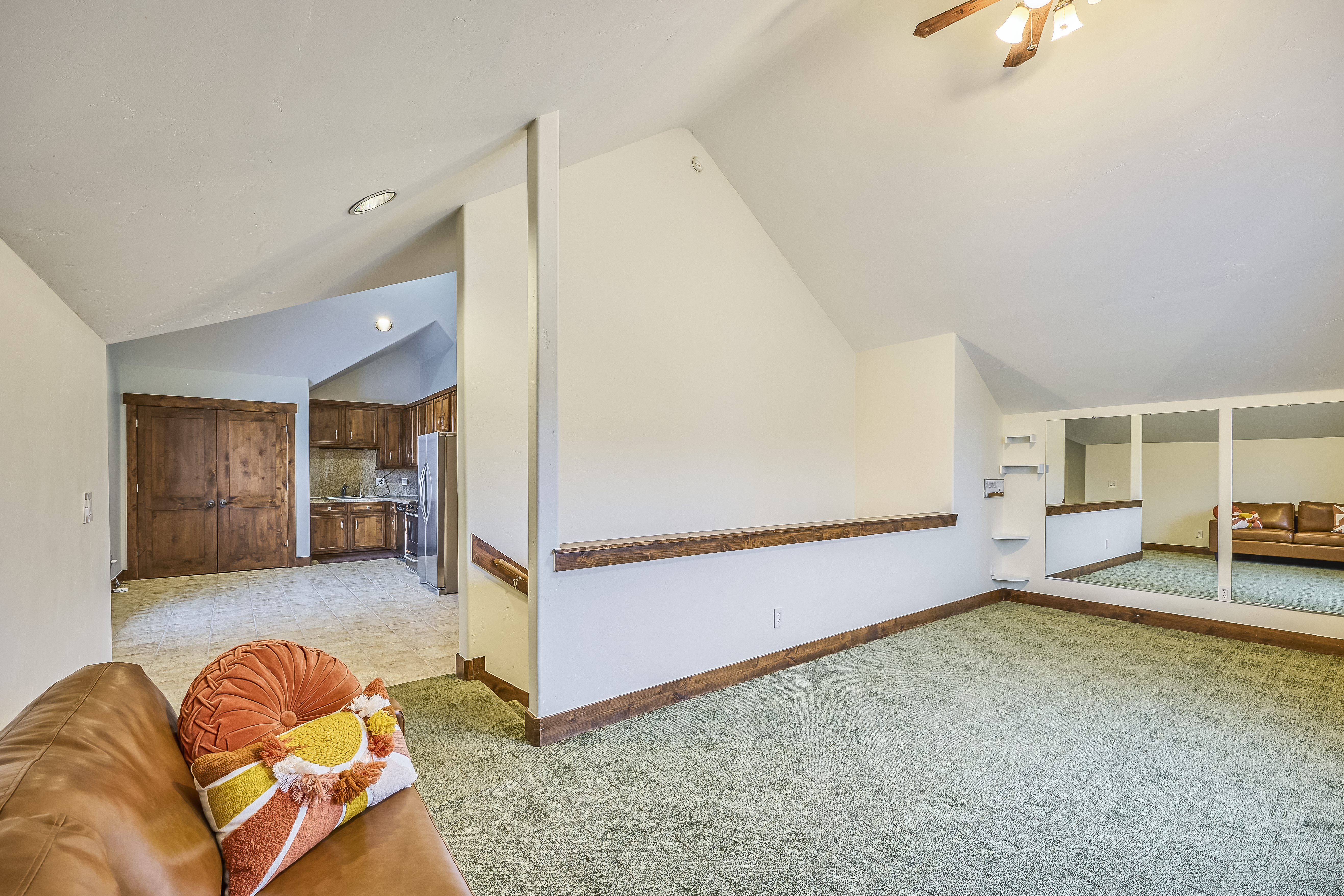 The living room is well-lit, featuring a plush green carpet and neutral-colored walls. A comfortable leather sofa with decorative cushions and a partial view into the kitchen space invites relaxation. A large mirror reflects the room, enhancing the sense of space and brightness.