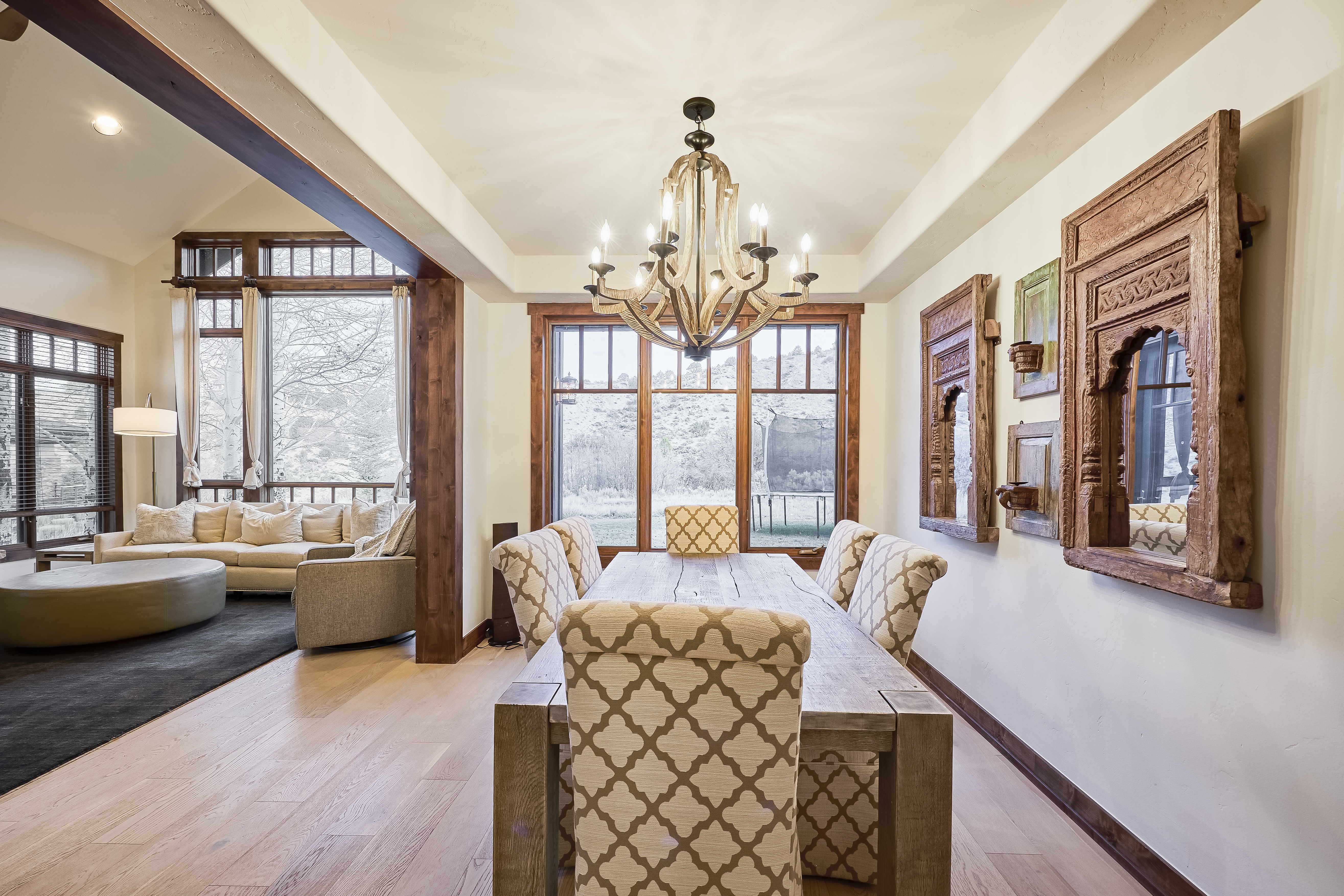 This is a bright dining room with hardwood floors, a long wooden table, and upholstered chairs. The room is illuminated by a chandelier and natural light from a large window overlooking a wintery landscape. Decorative wooden panels adorn the walls, adding character to the space.