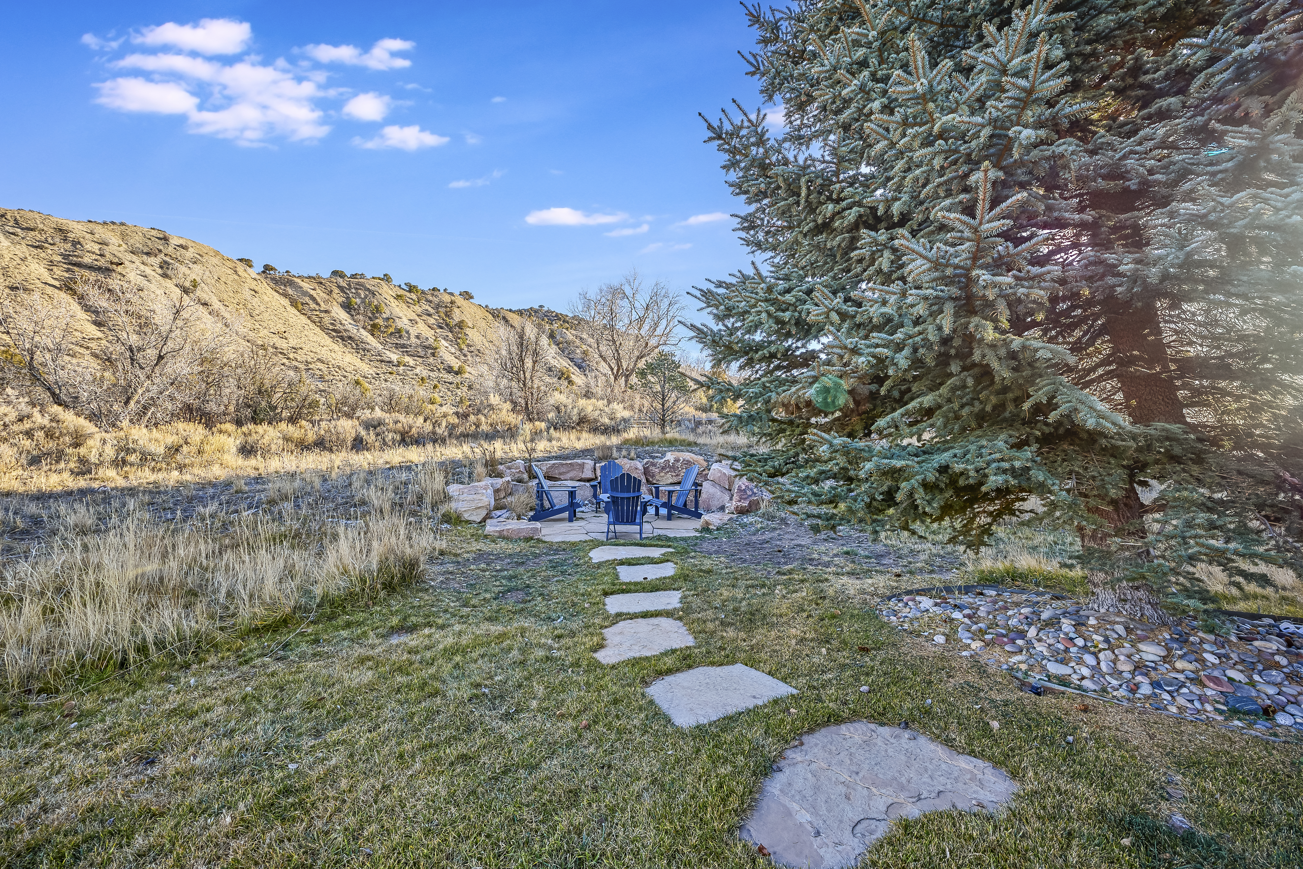 This image showcases a beautifully landscaped yard featuring a stone pathway leading to a cozy outdoor seating area with Adirondack chairs arranged around a fire pit. A mature evergreen tree anchors the scene, creating a sense of privacy and tranquility. The surrounding natural landscape and well-maintained lawn enhance the property's appeal.