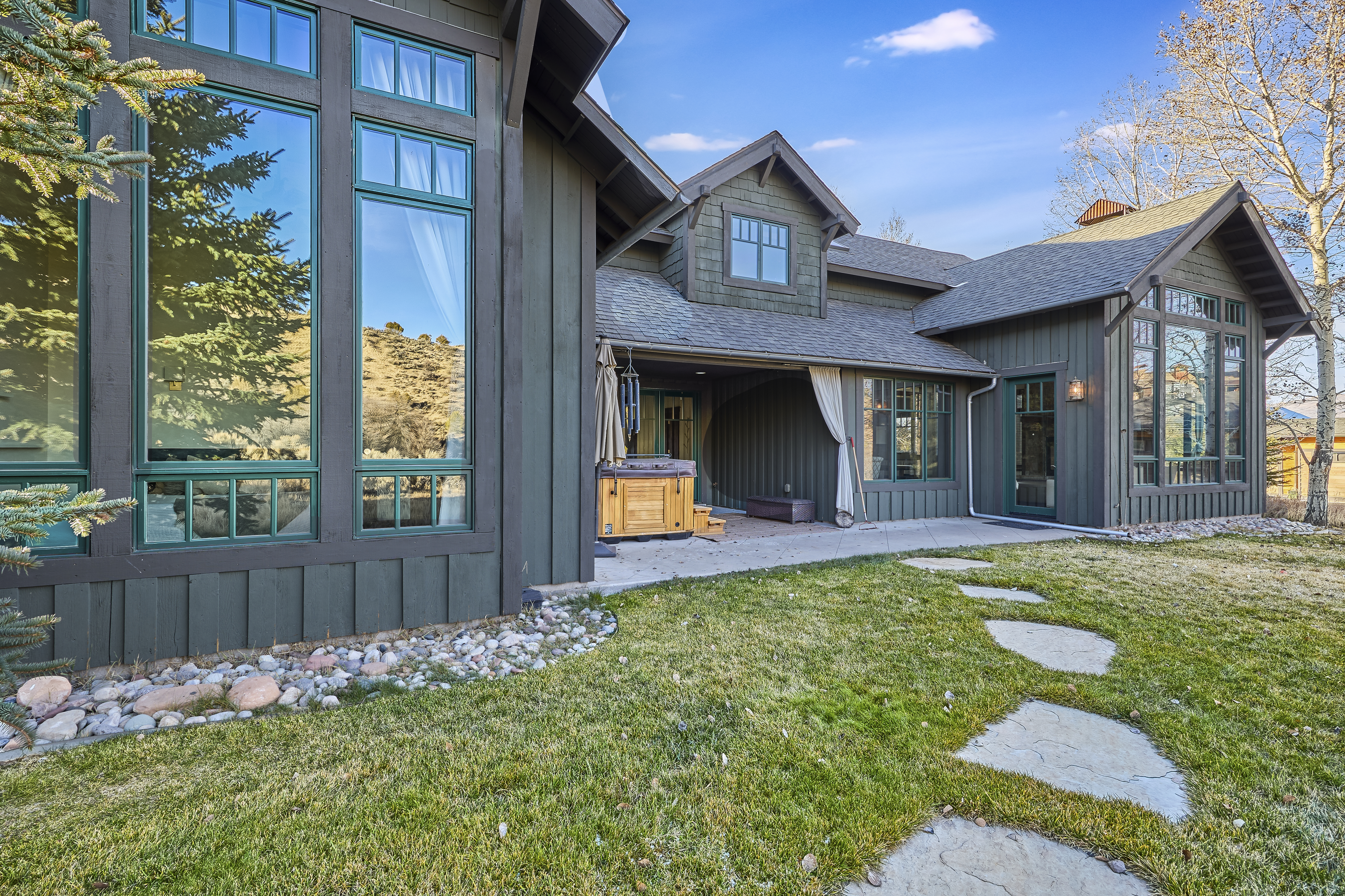 This is a rear view of a unique, wood-sided home featuring a patio, hot tub, and lush lawn with stone pavers. The architecture is characterized by large windows with green trim, a gabled roof design, and mature trees in the background. The image showcases an inviting outdoor living space perfect for relaxation and entertainment.