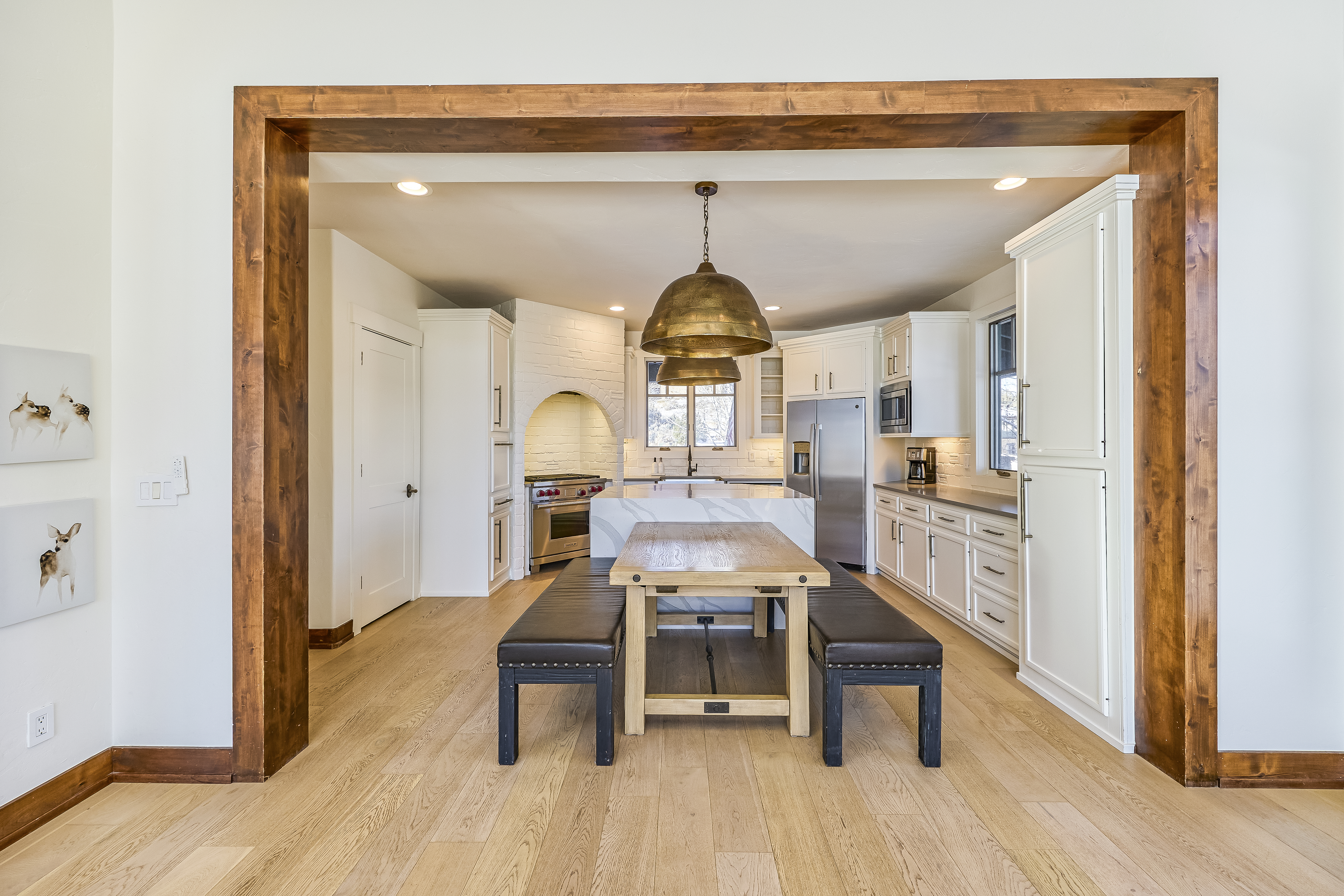 This is a bright, open kitchen featuring white cabinetry and countertops, stainless steel appliances, and a custom-built dining table area. Two large, domed pendant lights hang above the dining table. The kitchen is framed by a striking wooden architectural detail, providing an inviting and stylish transition from another room.