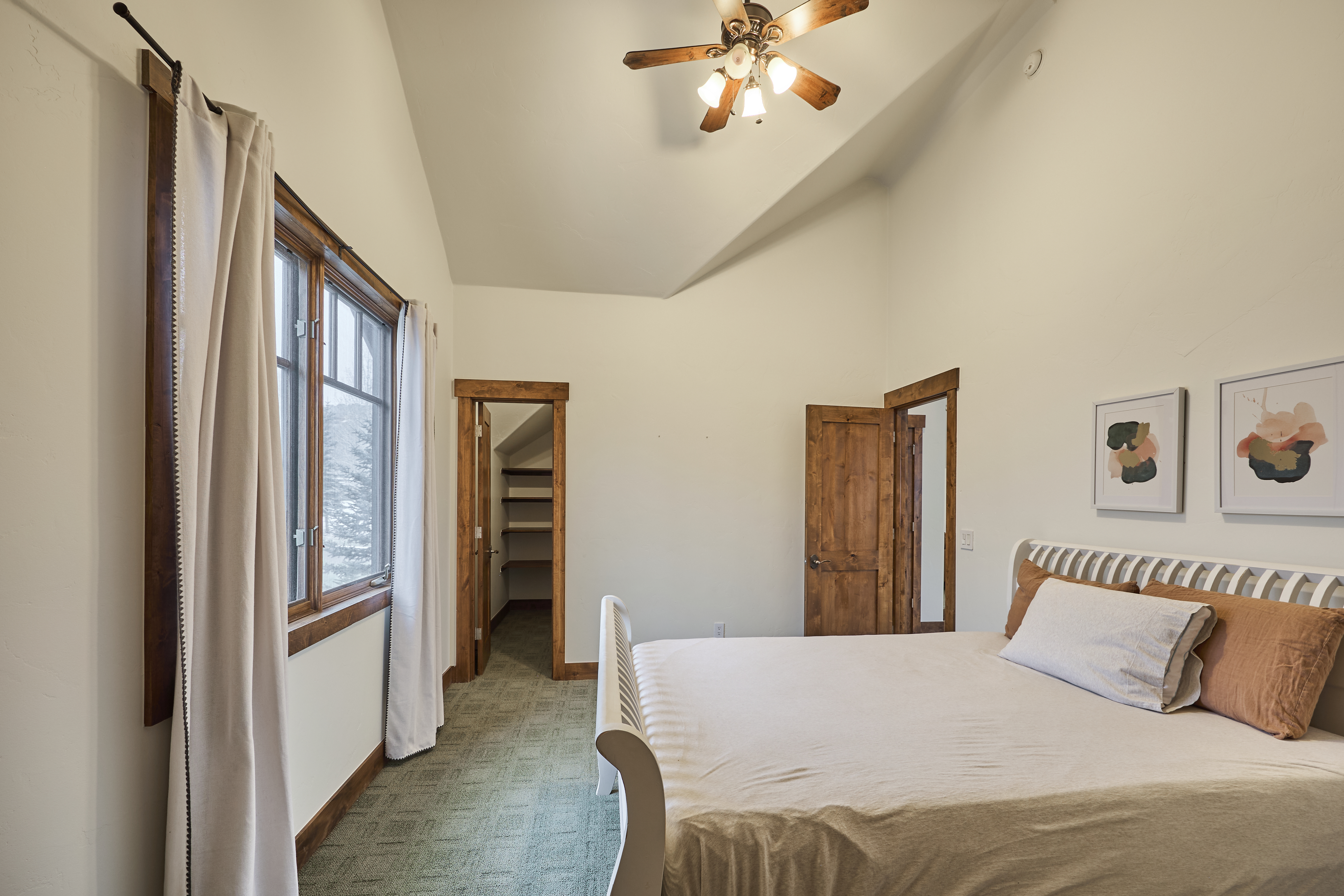 The image showcases a well-lit primary bedroom featuring a vaulted ceiling and rustic wood accents. A white sleigh bed with neutral linens takes center stage, complemented by artwork above the headboard. An open closet door and a glimpse through the window suggest a tranquil and inviting atmosphere.
