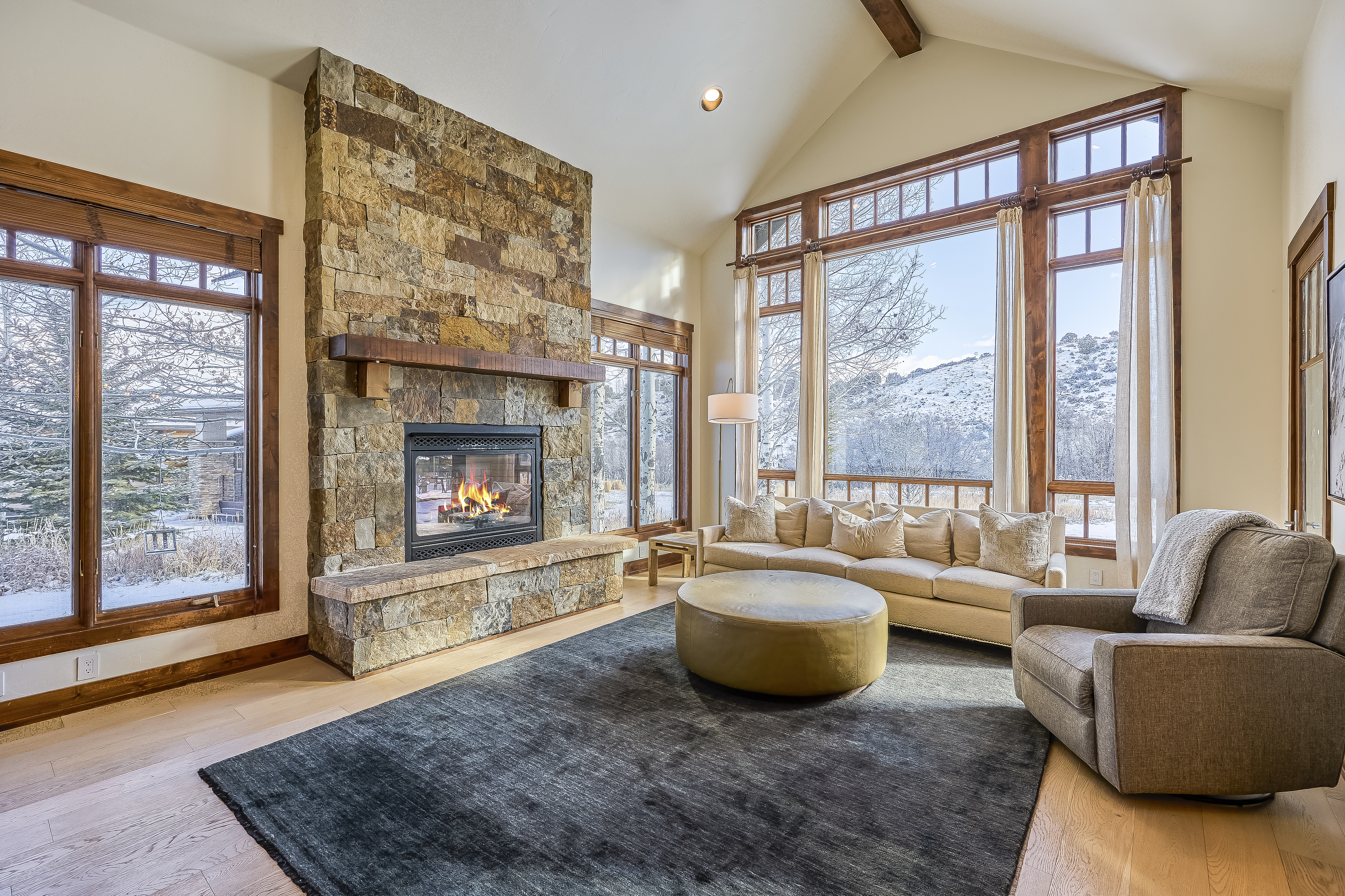 This inviting living room showcases a striking floor-to-ceiling stone fireplace, providing a warm focal point. Large windows frame scenic views, bathing the space in natural light and complementing the neutral color palette. The layout includes a comfortable sofa, armchair, and a round ottoman, creating a cozy and elegant atmosphere.