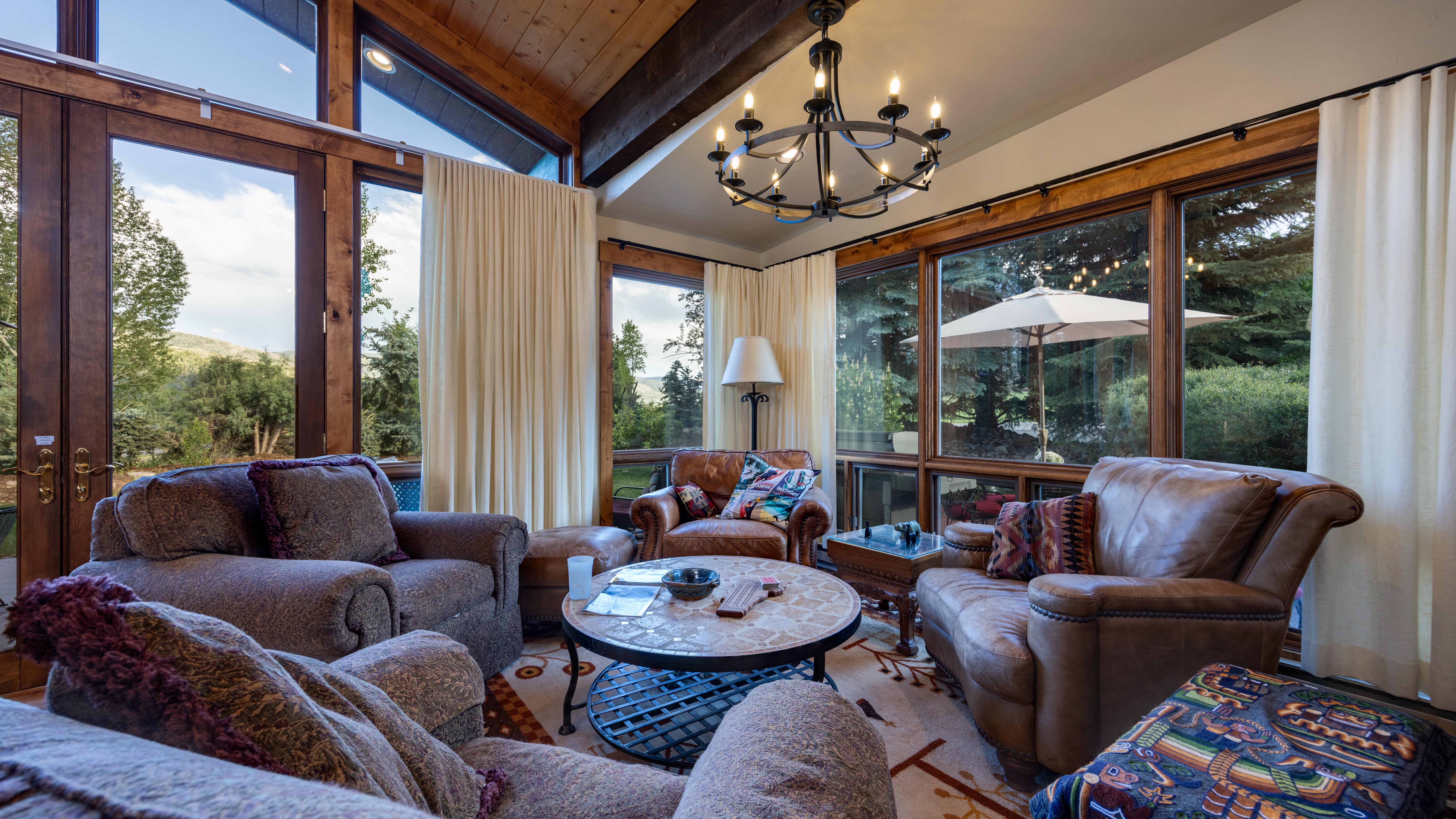 This cozy living room features a warm, rustic aesthetic with comfortable leather and upholstered seating arranged around a circular stone-topped coffee table. The space is anchored by a striking wrought-iron chandelier and large, wall-to-ceiling windows that provide abundant natural light and serene views of the wooded outdoors. High wooden ceilings and elegant, floor-length curtains add to the sophisticated yet welcoming atmosphere of this retreat-style home.