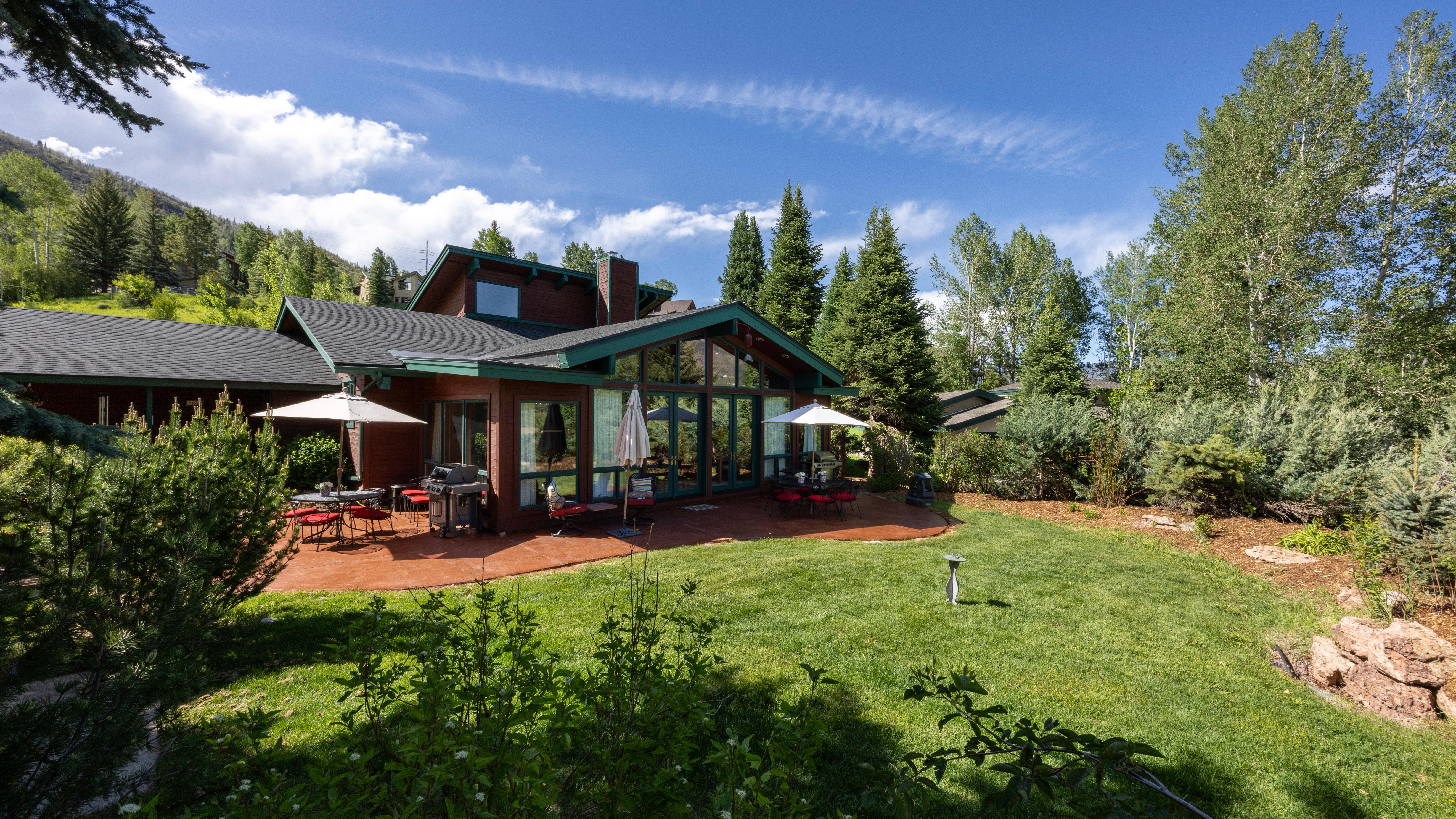 This rear view showcases a charming mountain-style home featuring a large, sun-drenched patio and a spacious, well-maintained lawn. The exterior boasts a warm wood siding complemented by vibrant green trim, while expansive floor-to-ceiling windows provide a seamless connection between the indoors and the scenic natural landscape. The property is surrounded by mature trees and lush greenery, creating a serene and private atmosphere ideal for outdoor dining and entertaining.