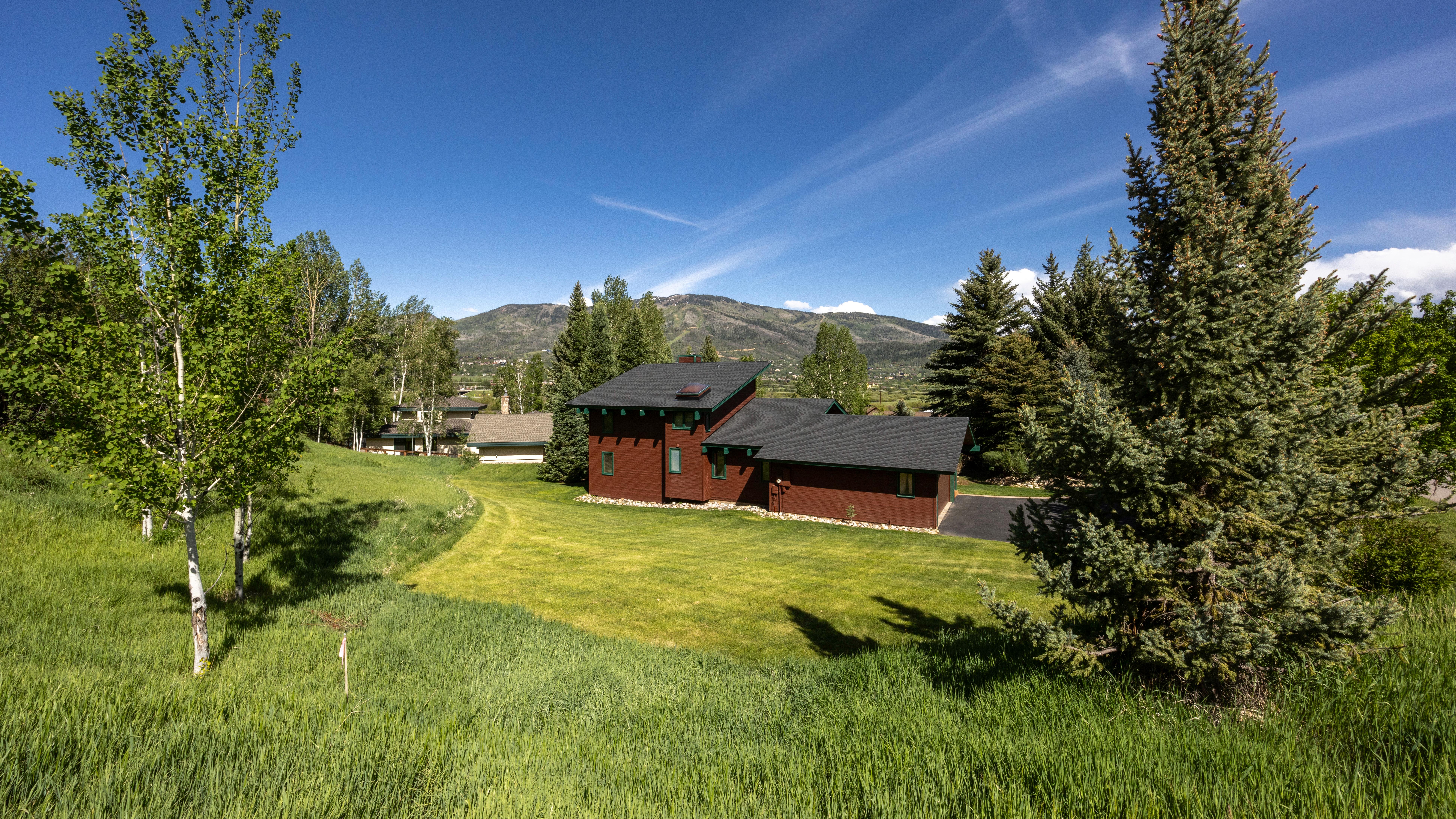 This stunning side view captures a dark brown, multi-level home nestled against a backdrop of scenic rolling hills and mountain views. The property features a manicured lawn, a paved driveway, and lush surrounding landscaping including aspen and pine trees. The home's design is modern-rustic, complementing the natural beauty of its quiet, scenic environment.