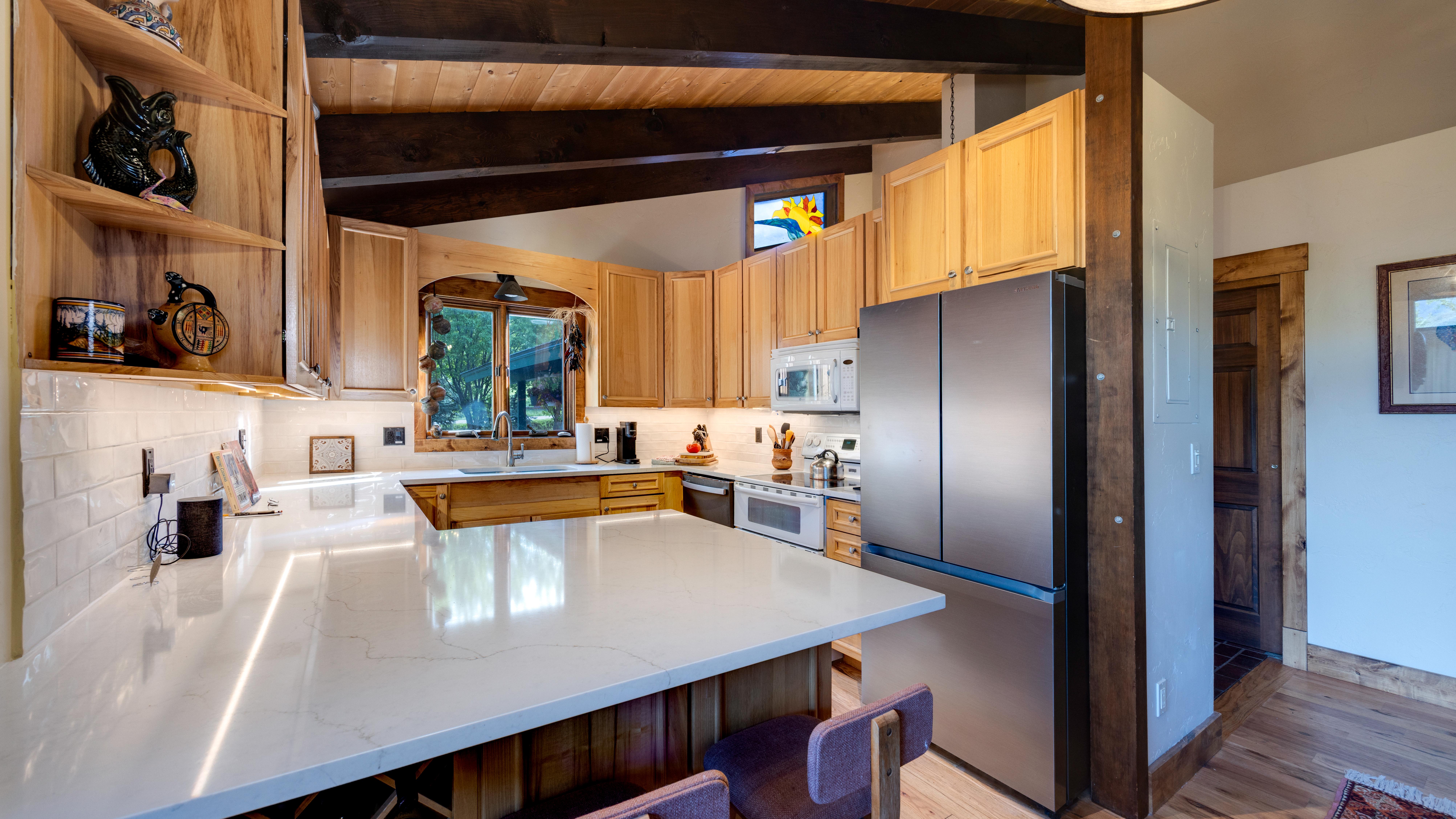 This rustic-inspired kitchen features warm wood cabinetry, a expansive white quartz countertop with breakfast bar seating, and stainless steel appliances. Exposed wooden ceiling beams and a charming stained-glass accent window add character to the space. The blend of farmhouse textures and modern convenience creates a cozy and inviting atmosphere.