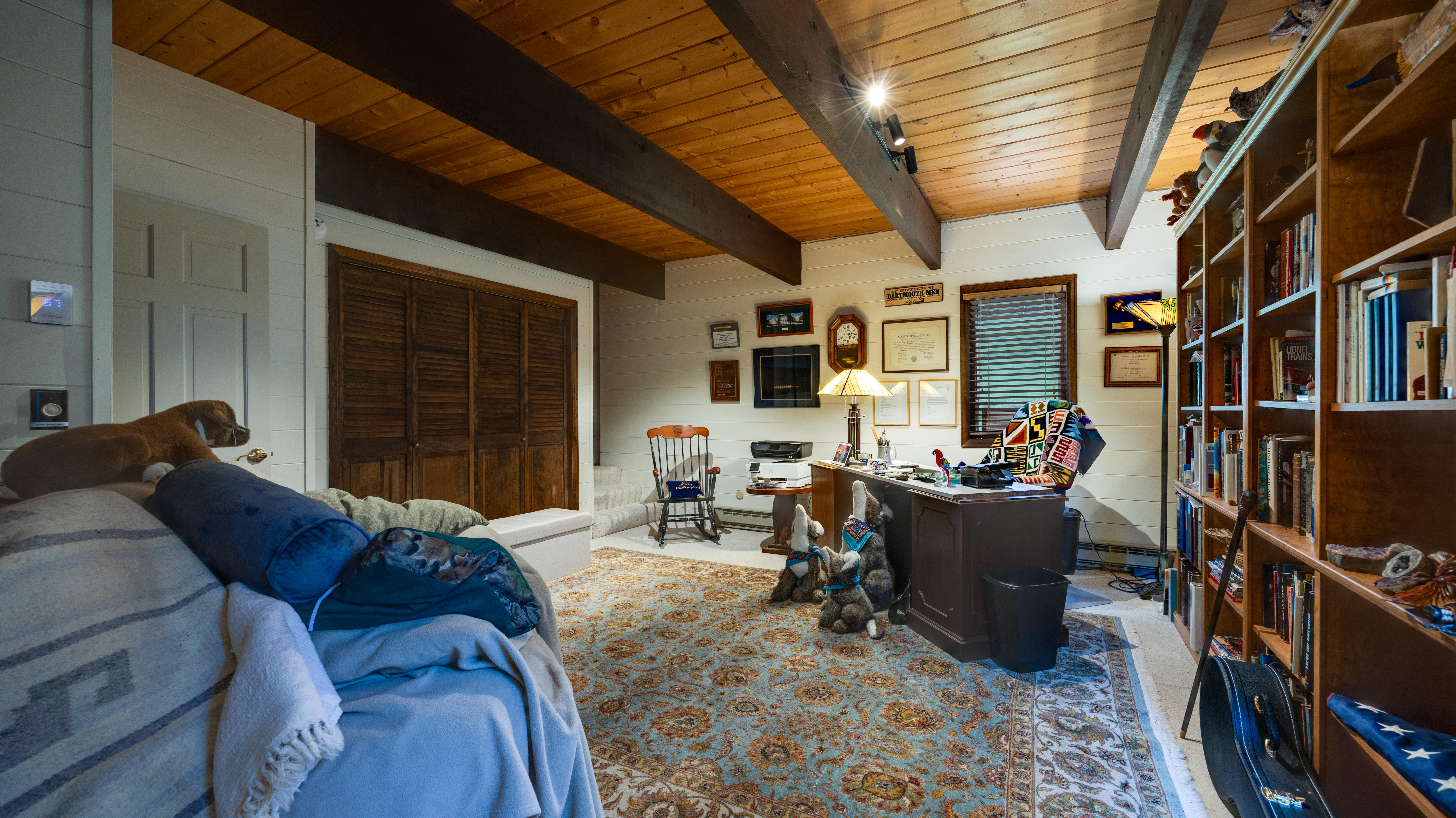 This cozy home office features warm wood-paneled ceilings with exposed beams and rustic white walls, creating a welcoming and studious atmosphere. The room is well-appointed with a substantial wooden desk, built-in shelving packed with books, and an ornate Persian-style rug that adds a touch of traditional elegance. A collection of small stuffed animals and personal décor items gives the space a lived-in, comfortable charm.