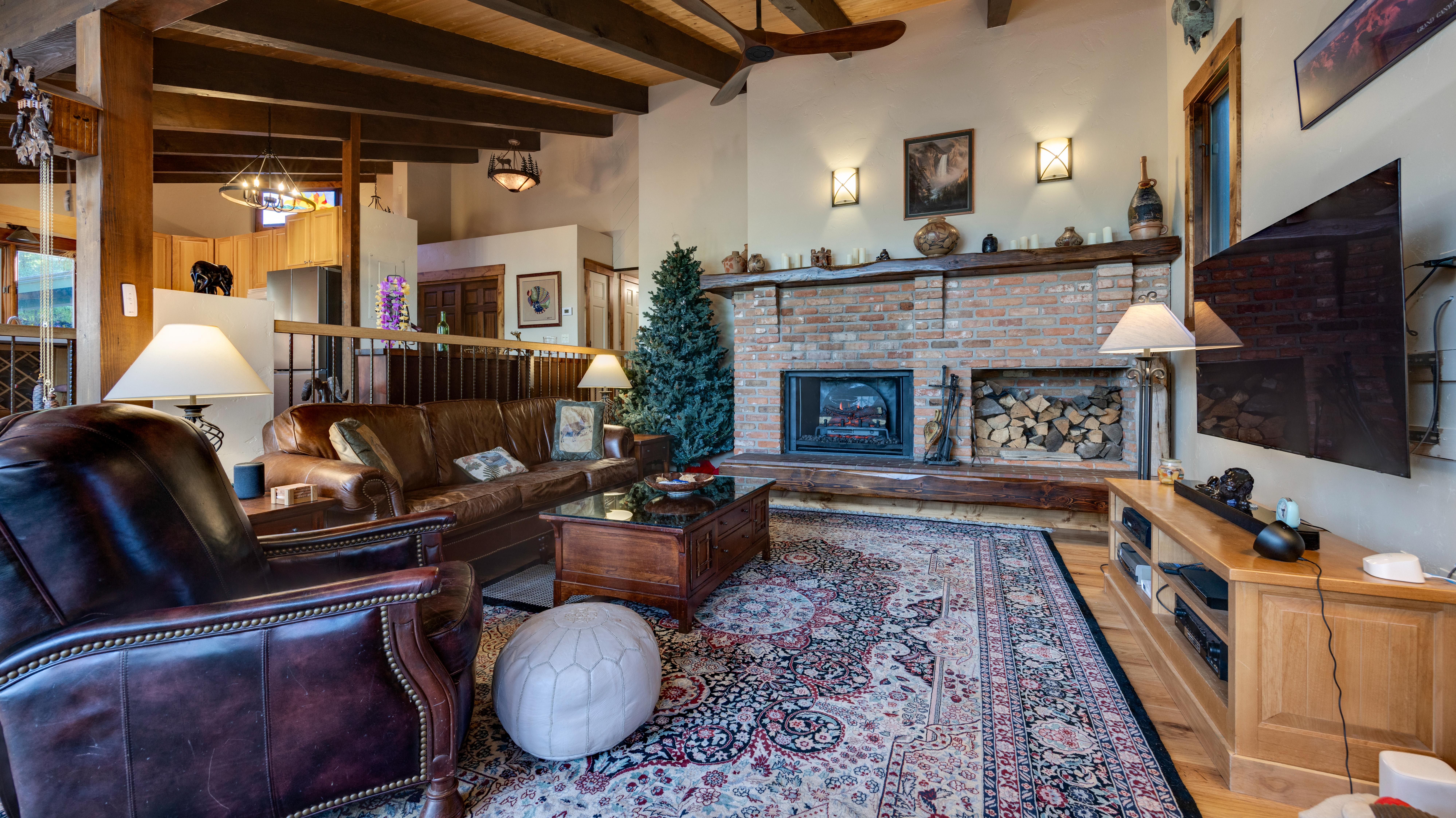 This cozy, rustic living room features dark leather furniture, an eye-catching brick fireplace with a wood mantle, and a large, ornate area rug. The room is accented by warm lighting, exposed wooden ceiling beams, and integrated shelving for media, creating a welcoming and comfortable atmosphere perfect for relaxation. A Christmas tree tucked near the fireplace adds a festive touch to this well-appointed space.