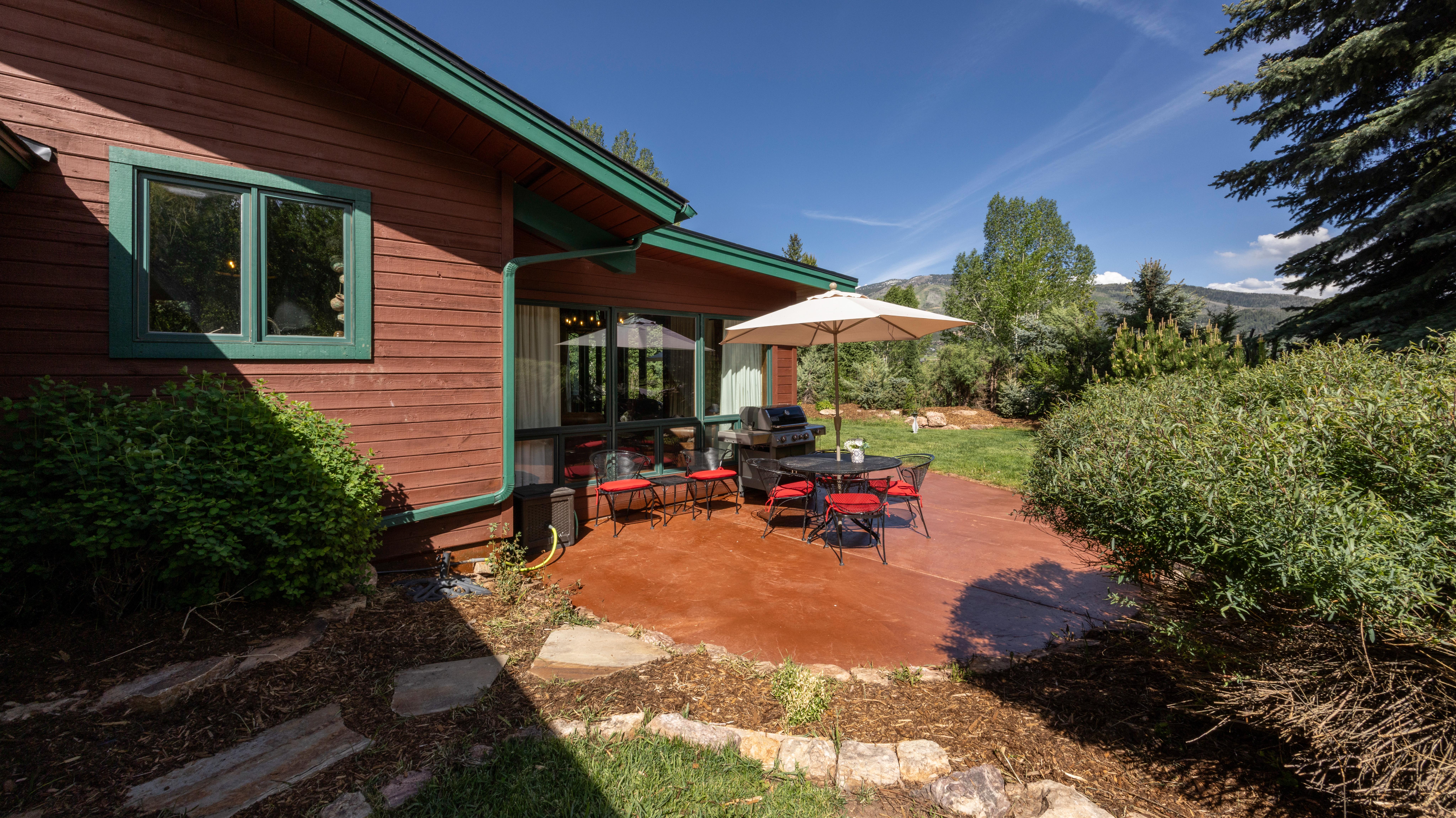 This outdoor living space features a spacious, reddish-brown stained patio equipped with a dining table, chairs with red cushions, and a large umbrella. The area directly abuts the house, which showcases wood siding with green trim and a large sliding glass door. The backyard setting is tranquil, surrounded by lush green shrubs and trees with partial mountain views in the distance.