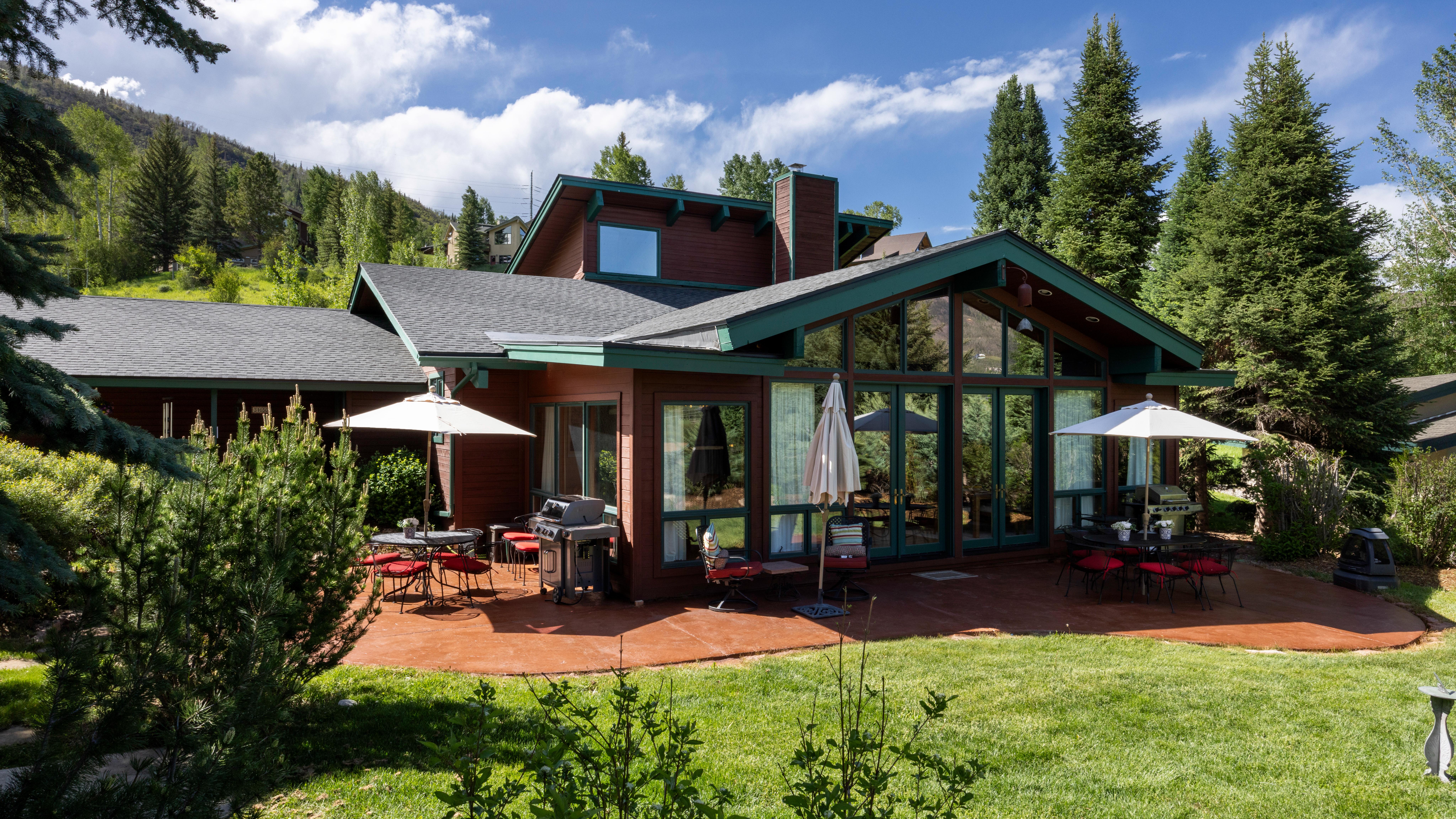 This rear view of a rustic mountain home highlights a spacious, stamped concrete patio perfect for outdoor dining and entertaining. The exterior features dark-toned wood siding, large floor-to-ceiling windows that allow for plenty of natural light, and green trim accents that blend into the surrounding pine forest and grassy landscape. The property provides a serene and welcoming atmosphere, ideal for enjoying the picturesque mountain setting.